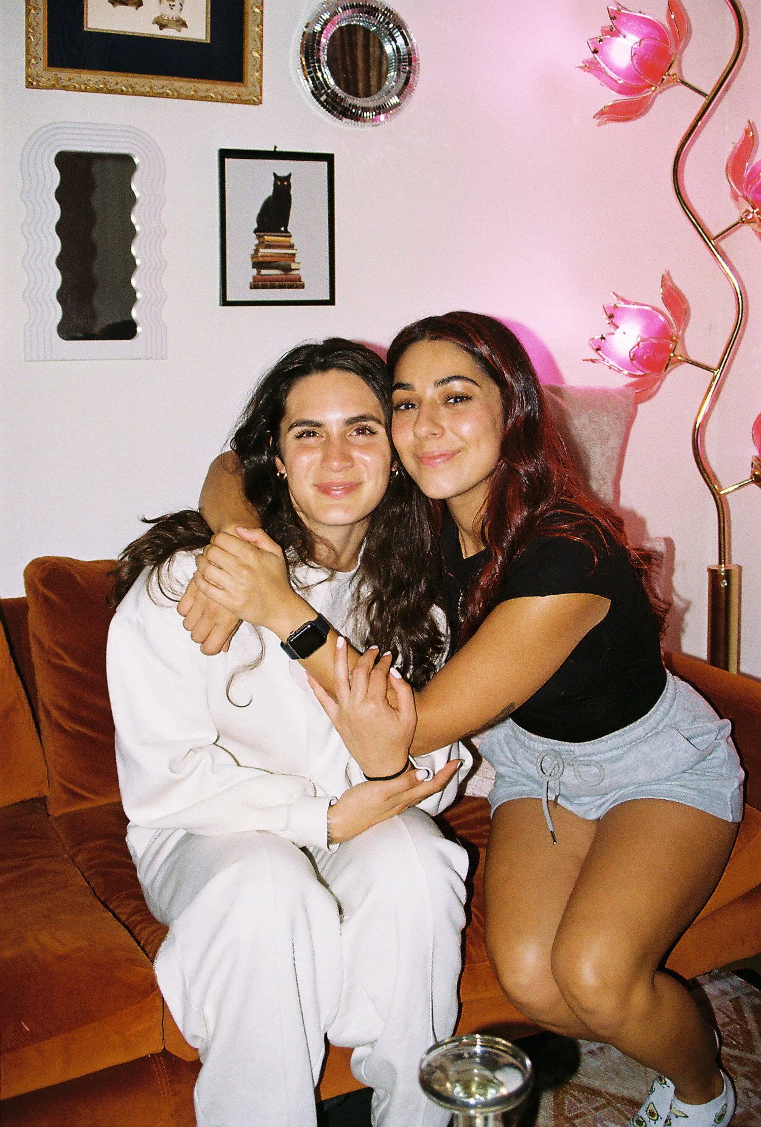 Two women sitting on a brown couch, smiling and hugging each other in a cozy, decorated room with pink walls, wall art, a mirror, and a pink flower-shaped floor lamp.
