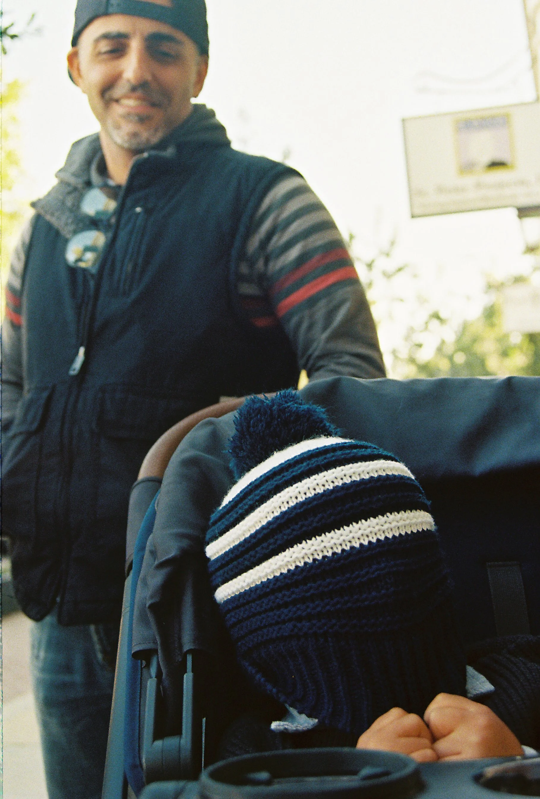 A man wearing a black cap and vest smiling at a camera, with a child in a stroller wearing a striped knit hat.
