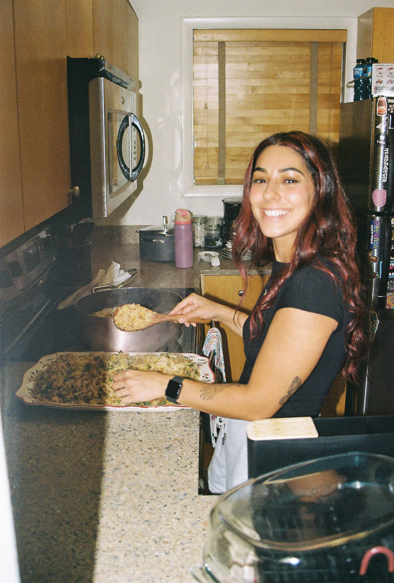 A woman with long, wavy red hair cooking rice or a similar dish in a black skillet on a kitchen stove, smiling at the camera, with a tray of cooked food in front of her in a kitchen setting.