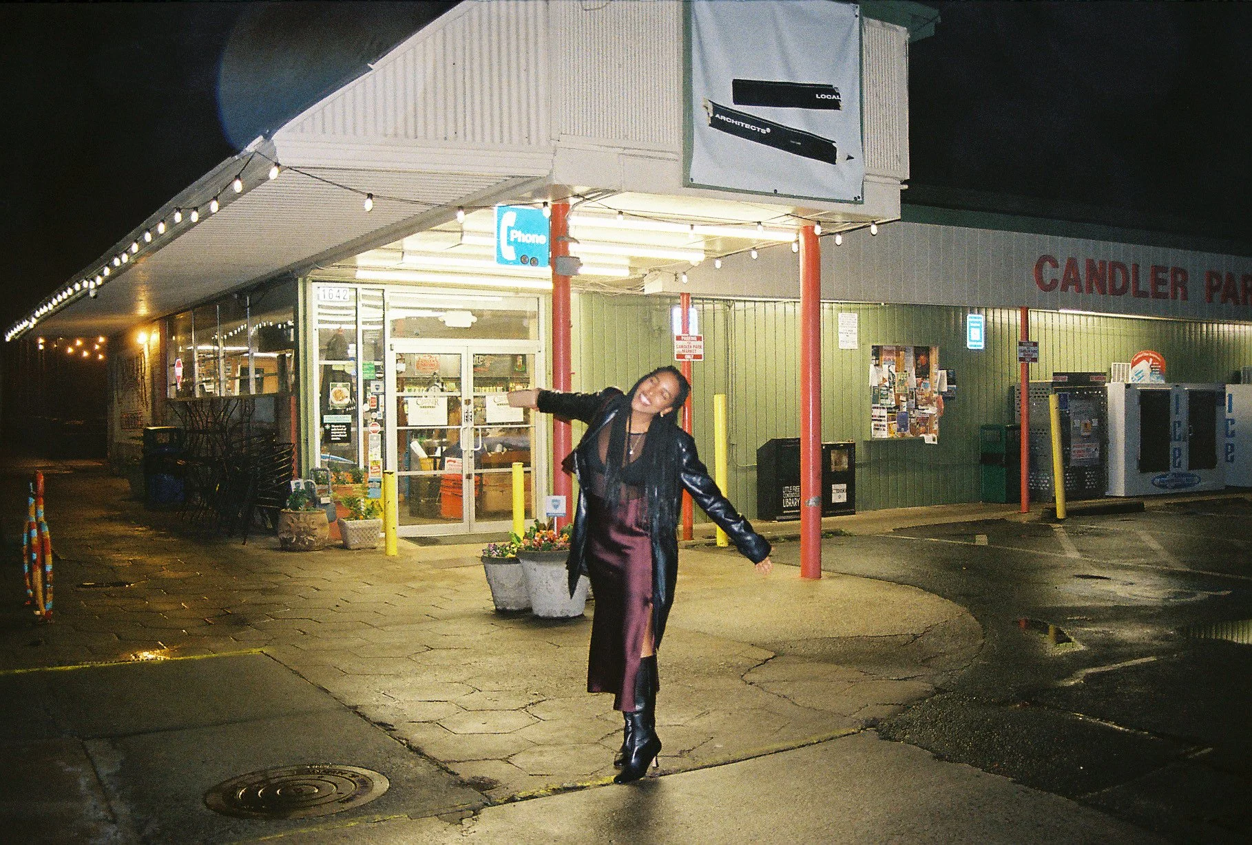 A woman in a black leather jacket, purple dress, and high boots dancing in front of a grocery store at night, with wet pavement reflecting light.