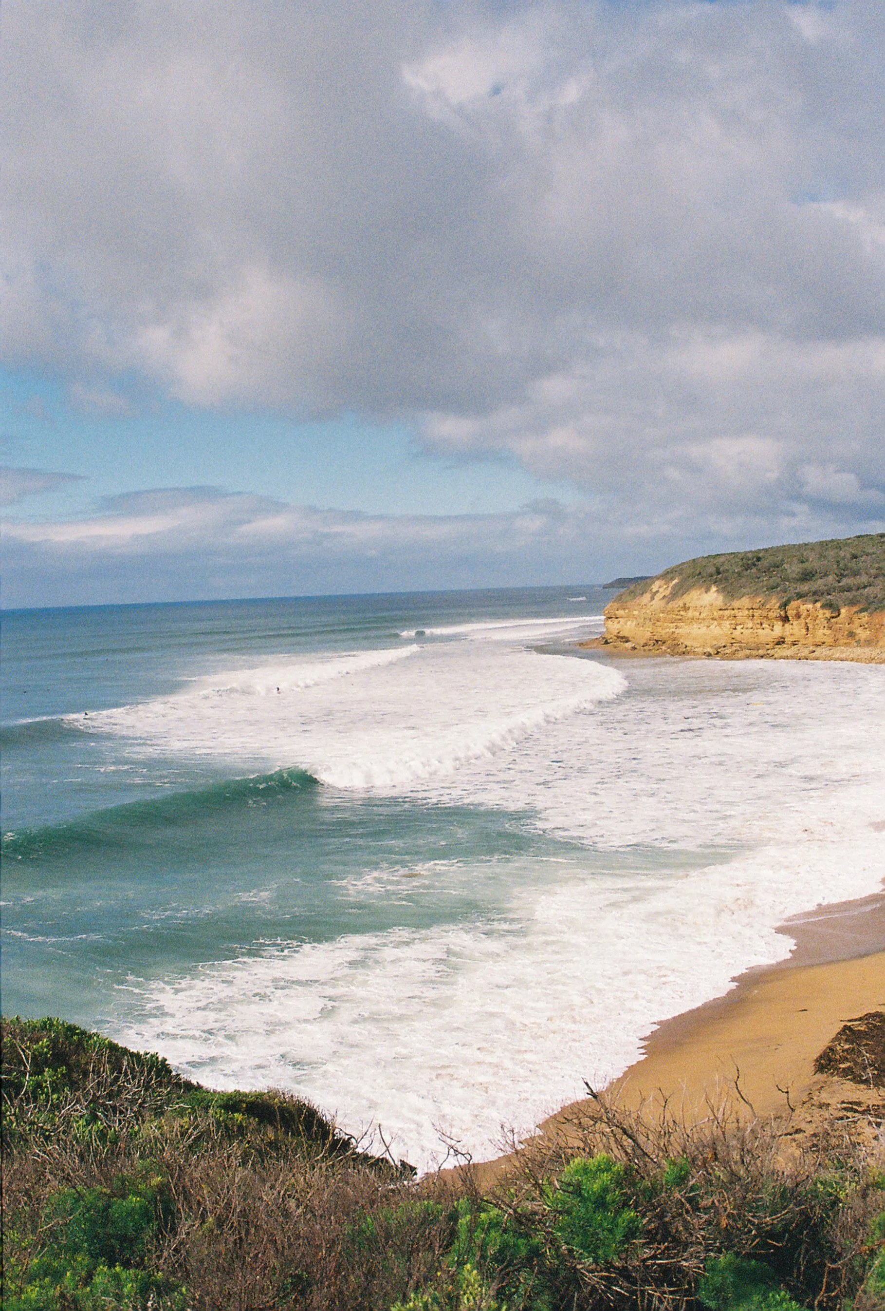A view of a coastline with waves hitting the shore, a rocky cliff on the right, and a cloudy sky overhead.