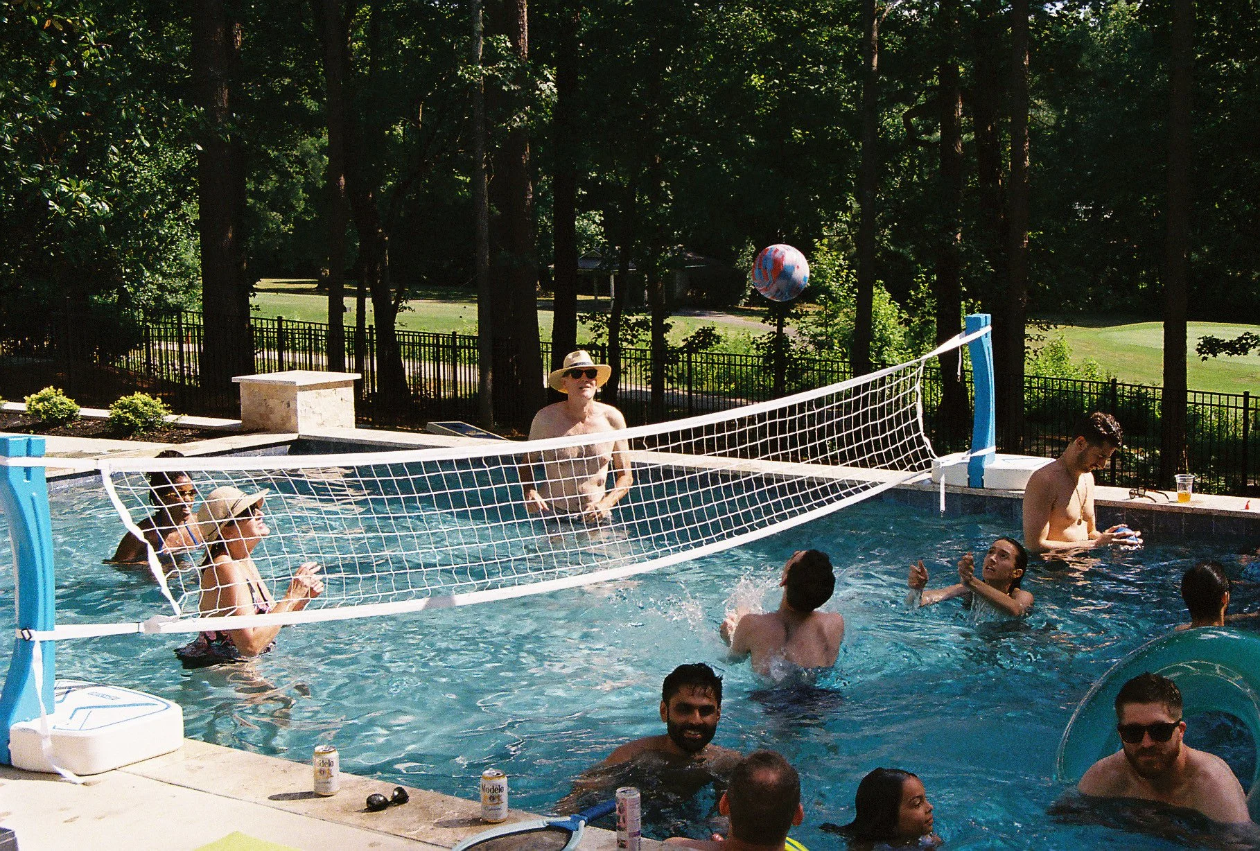 People swimming and playing volleyball in a backyard pool on a sunny day, with trees and a golf course in the background.