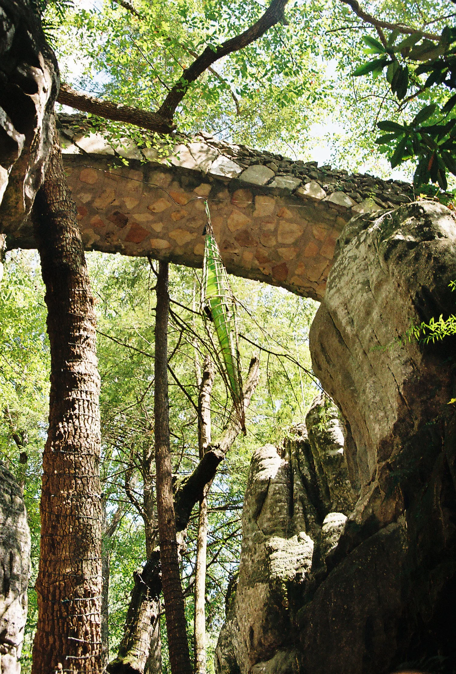 View of a stone bridge arching over a rocky and wooded area with tall trees and green foliage.