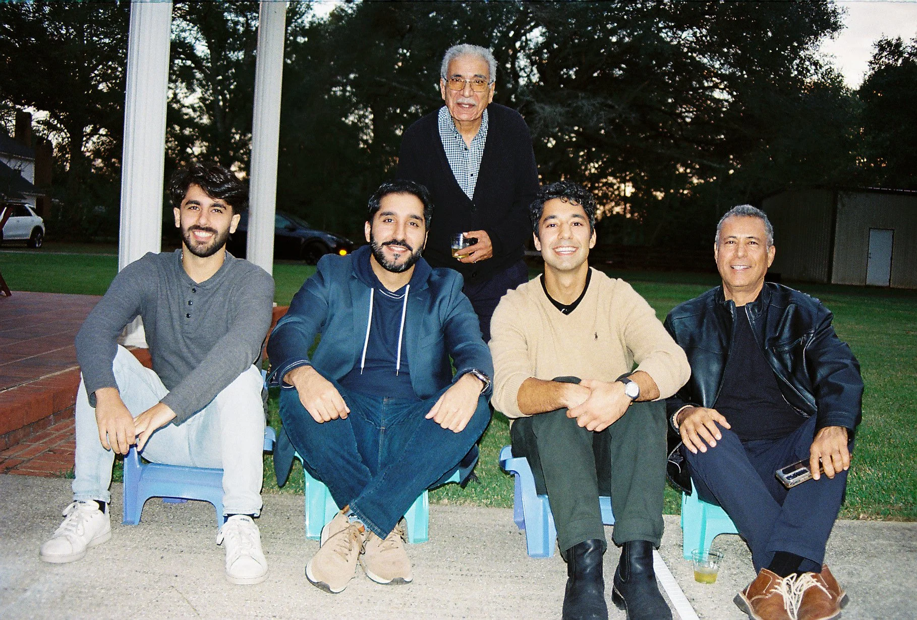 Five men sitting outdoors on small chairs, smiling at the camera, with trees and a building in the background during sunset, one standing behind them holding a drink.