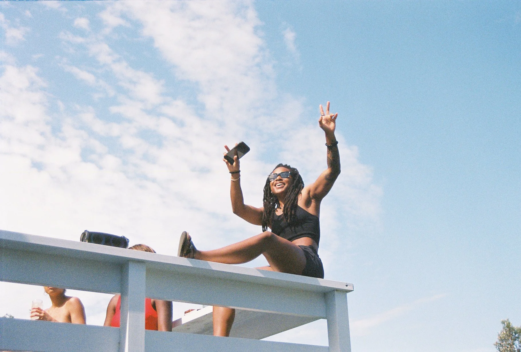 Young woman with dreadlocks, wearing sunglasses and black tank top, sitting on a roof with her legs outstretched, smiling and making peace signs with her hands, under a blue sky with white clouds.