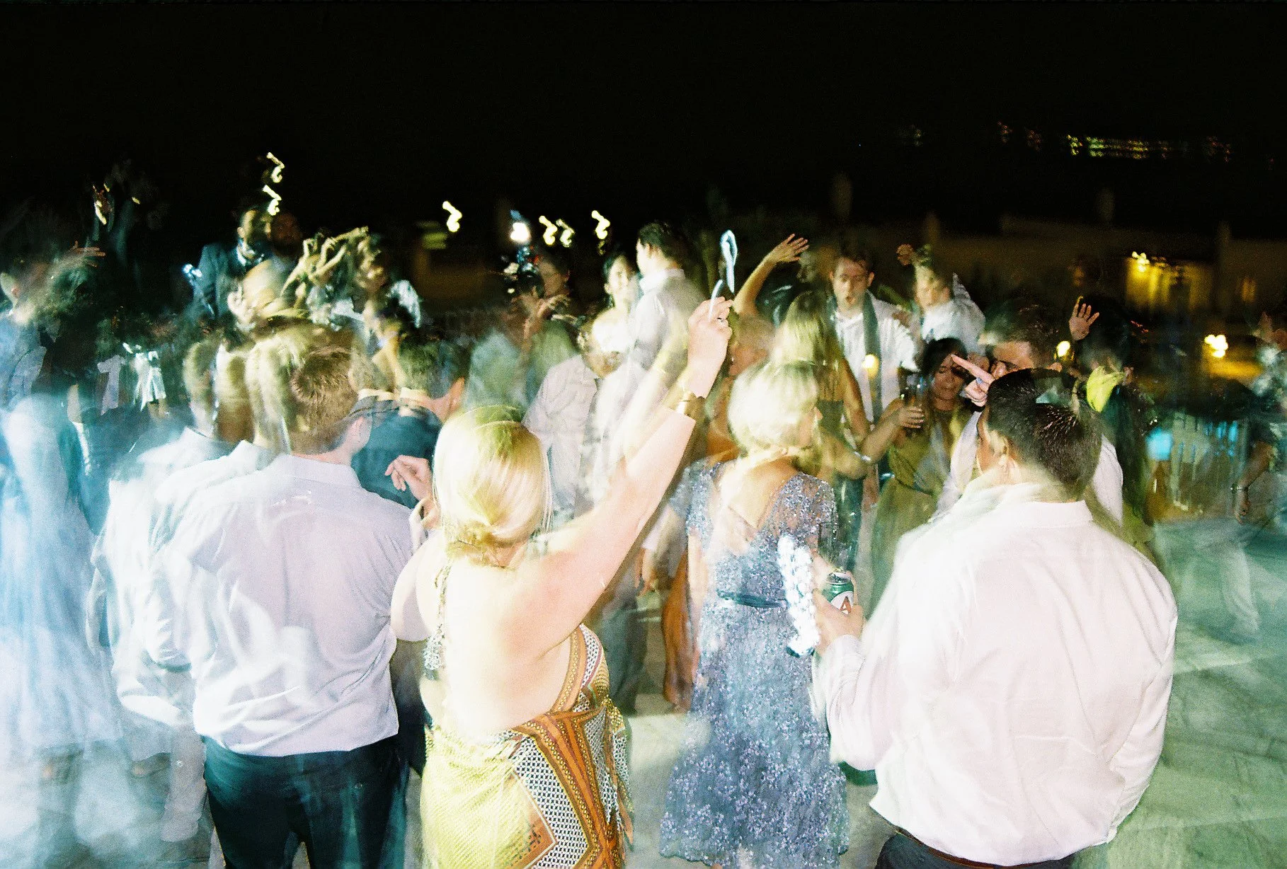Nighttime celebration with many people dancing and socializing, some holding drinks, blurry motion indicating dancing and lively atmosphere.