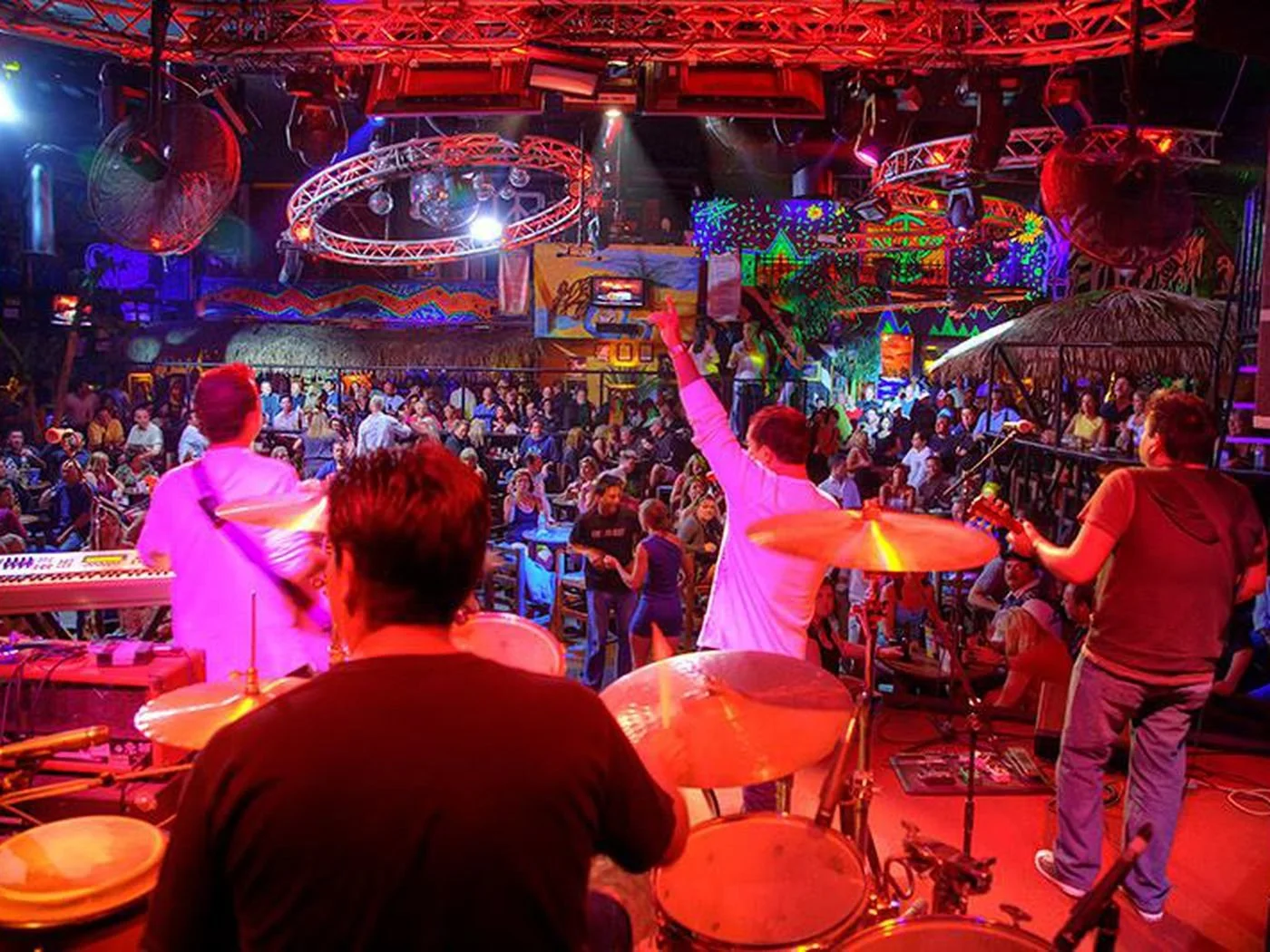 A photo of the view from the stage looking from behind the drummers shoulder. The singer has his arms in the air and you can see a couple dancing surrounded by other dancers on the dance floor of Cabo Wabo ! 