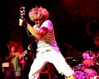 A photo od Sammy Stage with a Cabo Wabo Cantina t-shirt on rocking on his guitar. You can see people grooming in the background. Sammy has blonde curly hair is wearing all white with round sunglasses on. 