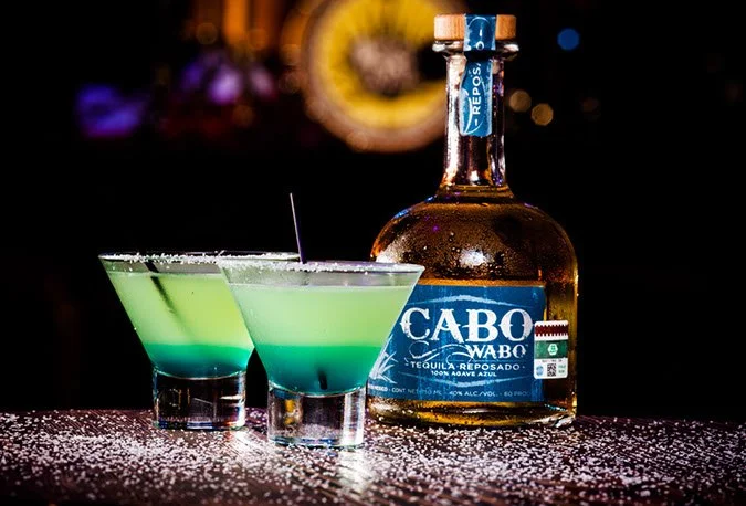 A photo of Cabo Wabo Cantina's tequila brand and a signature cocktail their make with it. There are two cocktails that have salted rims and are greener at the top and more turquoise at the bottom. There is the bottom of Cabo Wabo brand tequila sitti.