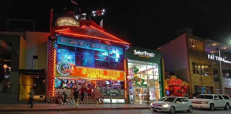 The outside of the El Squid row from the street. People are walking up to join the party. You can see their Neon sign and big logo. 