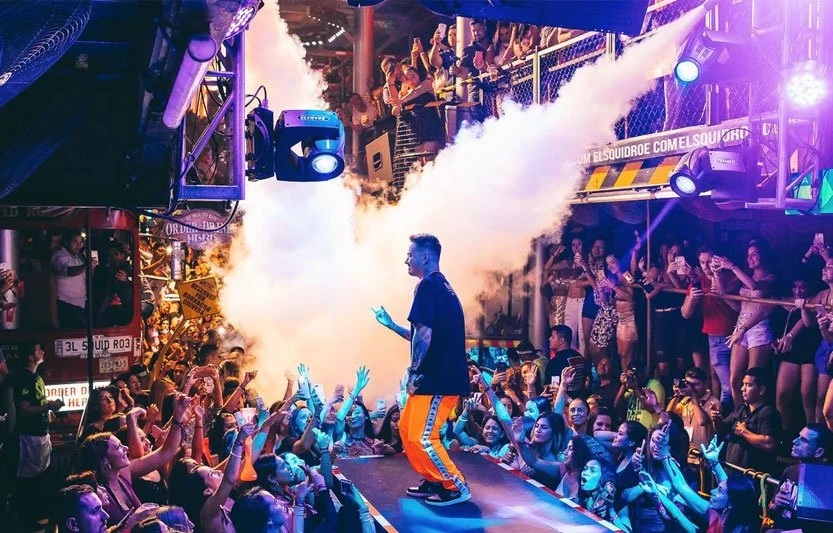 A photo of a DJ or dancer on stage in bright orange pants. There is smoke shooting down form the ceiling and the crowd has hitter hands and phones in the air.