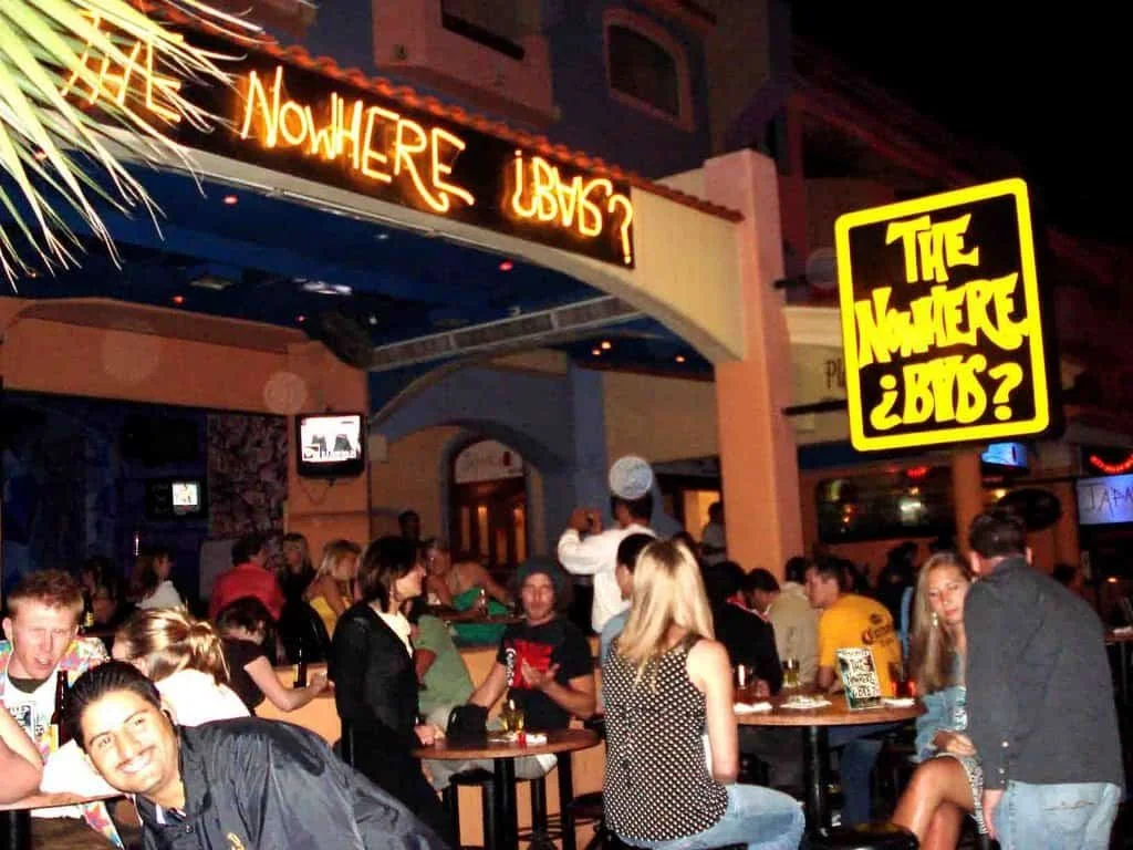 A photo outside of the Nowhere bar at night. With the neon sign and people sitting at high top tables outside. 