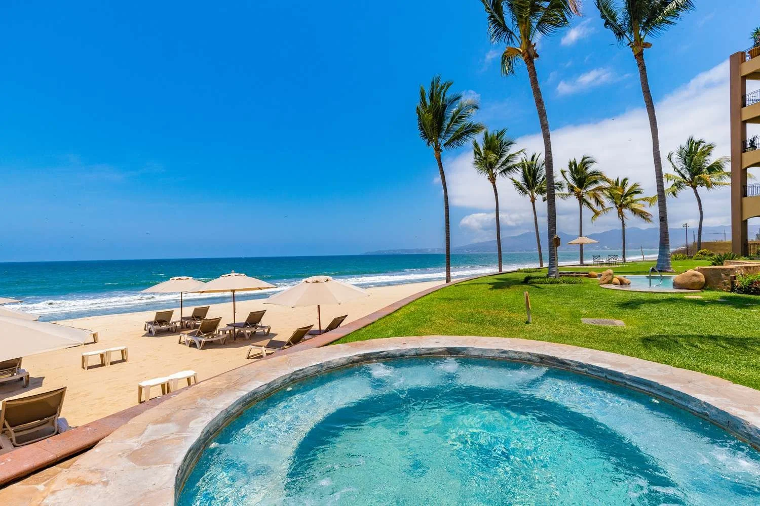A photo of one of the numerous hot tubs at villa la estancia resort and spa. The jacuzzi is overlooking the pristine white sandy beach and the teal blue sparkling ocean. There are palm trees and comfy loungers with umbrellas for shade in rows.