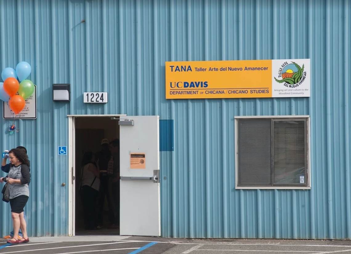 A teal metal building with an open door, a woman with curly hair drinking from a water bottle in the foreground, a balloon decoration, institutional signage, and a window.