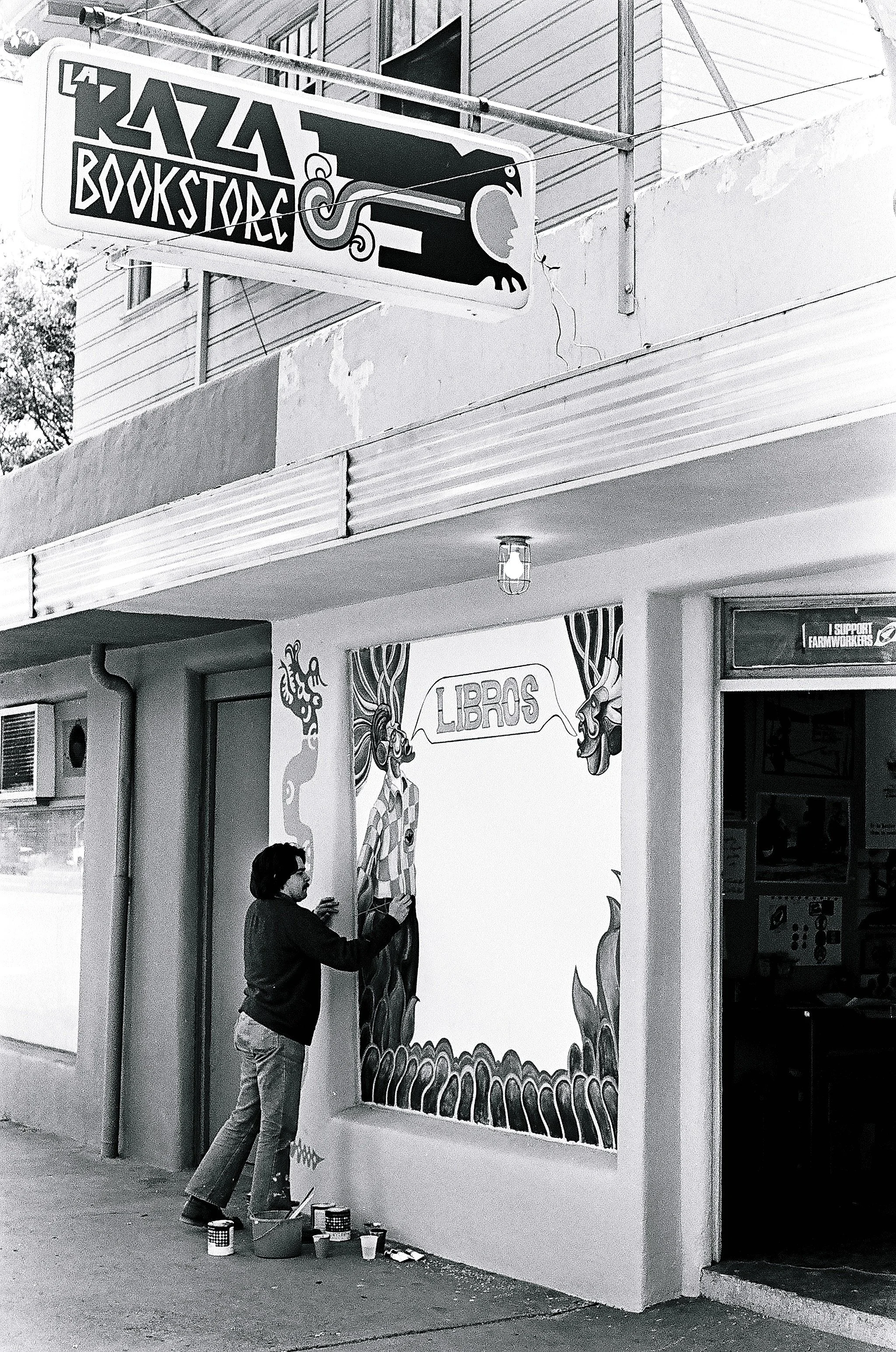 A person painting a mural on a wall, with the word 'LIBROS' inside a speech bubble and decorated with cartoon-like characters, near the entrance of a bookstore.