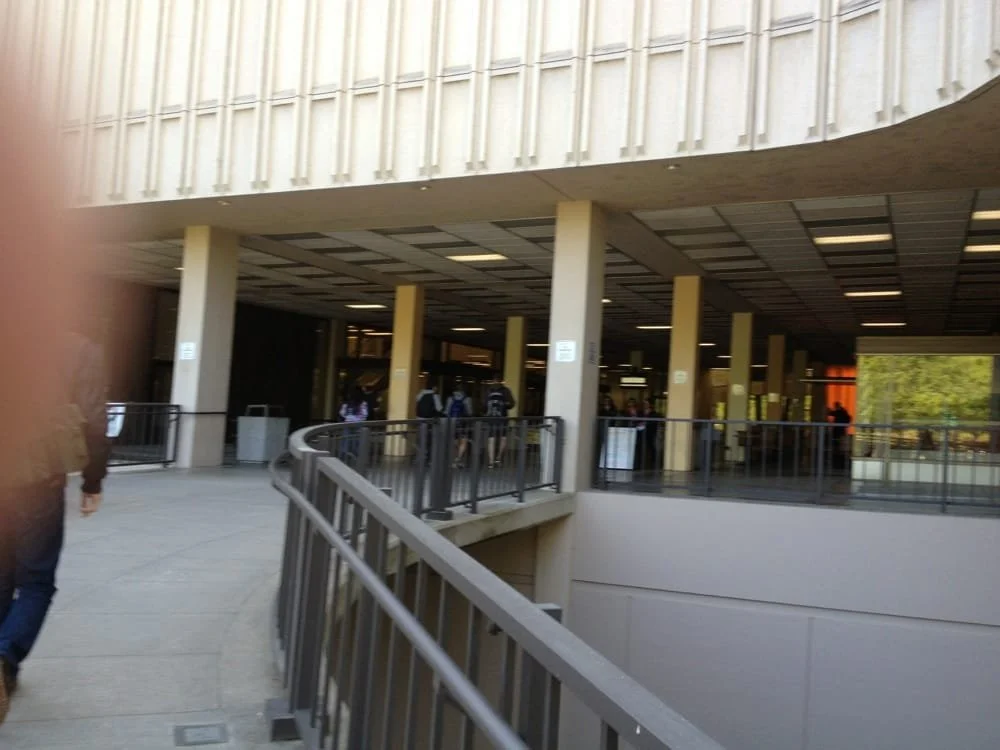 People walking into a modern building with a curved staircase and railing, tall columns, and large windows, possibly a public or institutional space.