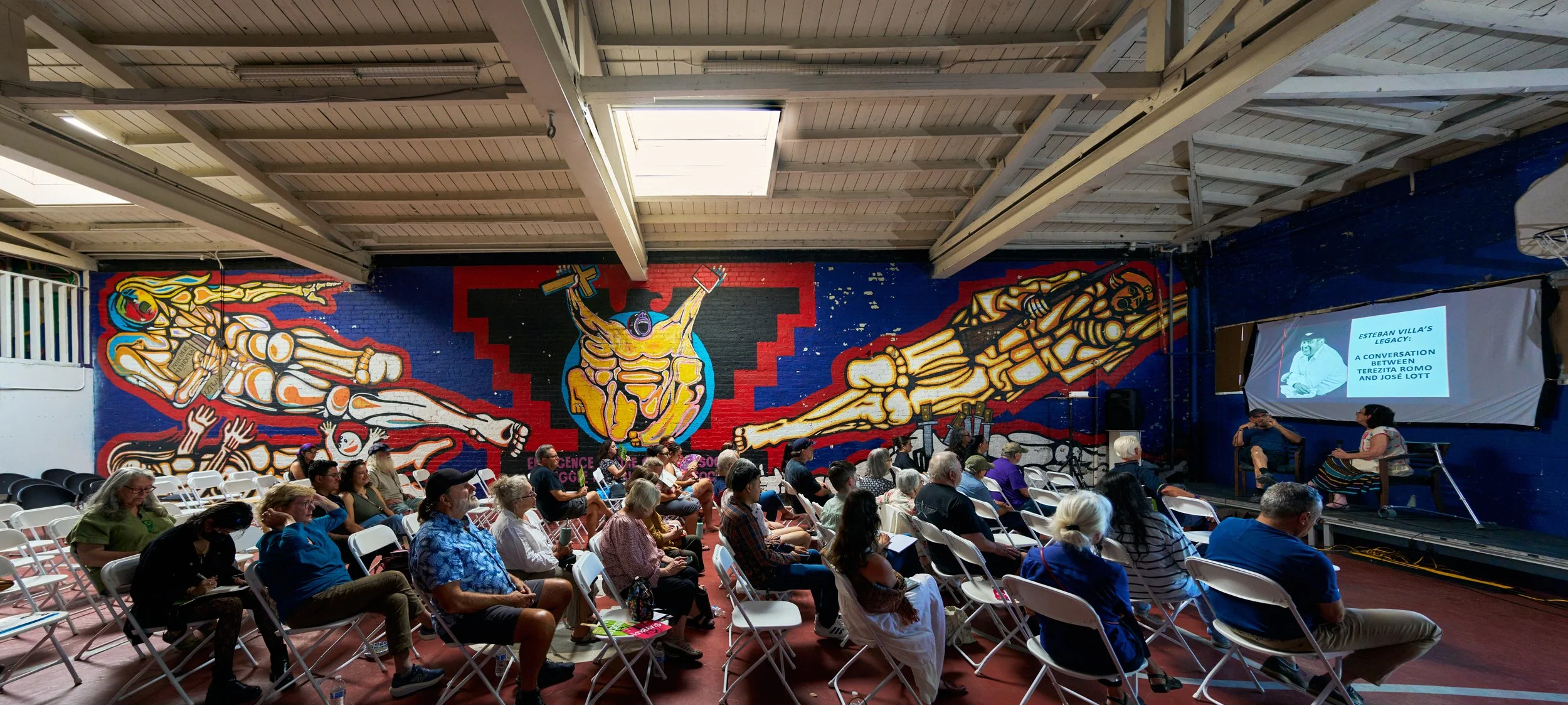 Audience seated in a hall watching a presentation, with a large colorful mural on the wall behind them depicting abstract figures and a central figure with raised arms. A projection screen on the right displays the title 'Esteban Villa's Legacy: A Conversation Between Terezita Romo And Jose Lott'.