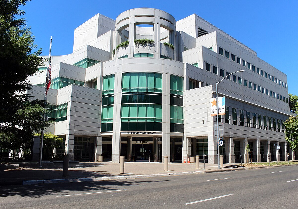 Modern multi-story building with glass windows and a white exterior, labeled California State Library, located on a city street under a clear blue sky.