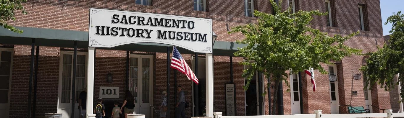 Entrance to the Sacramento History Museum, a brick building with a white sign that reads 'Sacramento History Museum.' People are entering the building, and there are trees and American flags outside.