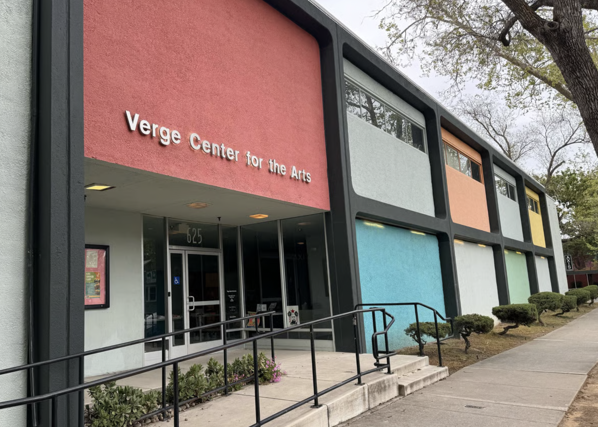 The exterior of a building labeled 'Verge Center for the Arts' with a colorful wall design on the upper facade, showing multiple rectangular sections painted in pastel colors, with bushes and a sidewalk in front.