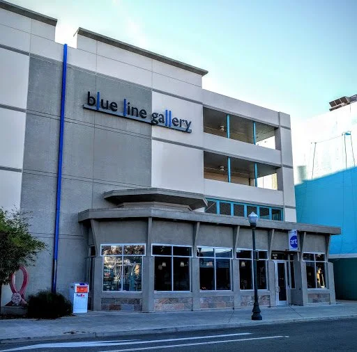 Exterior of a modern three-story building with a sign that reads "blue line gallery" on the upper level. The ground floor has large glass windows and a blue art sign near the entrance.
