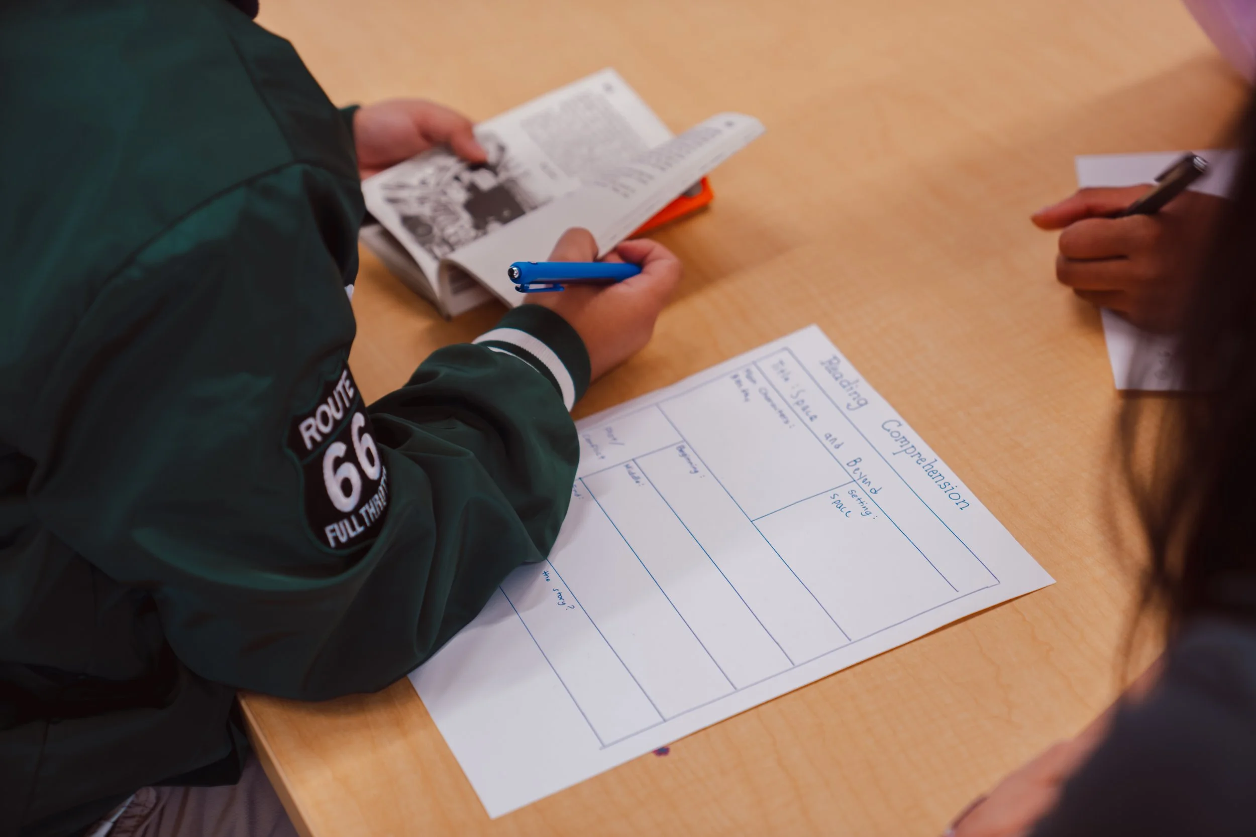 Two people sitting at a wooden table, one holding a magazine and a blue pen, and the other taking notes on a sheet of paper titled 'Reading Comprehension' with questions about reading and understanding.