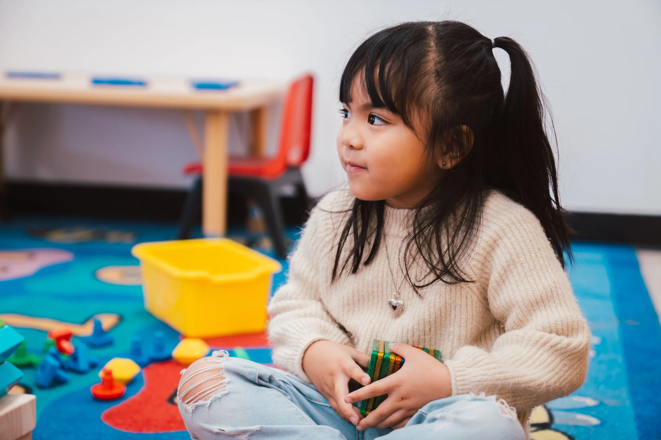 Young girl with dark hair in a ponytail sitting on a colorful play mat, holding small colorful building blocks, with toys and a yellow container on the mat, in a classroom or playroom setting.