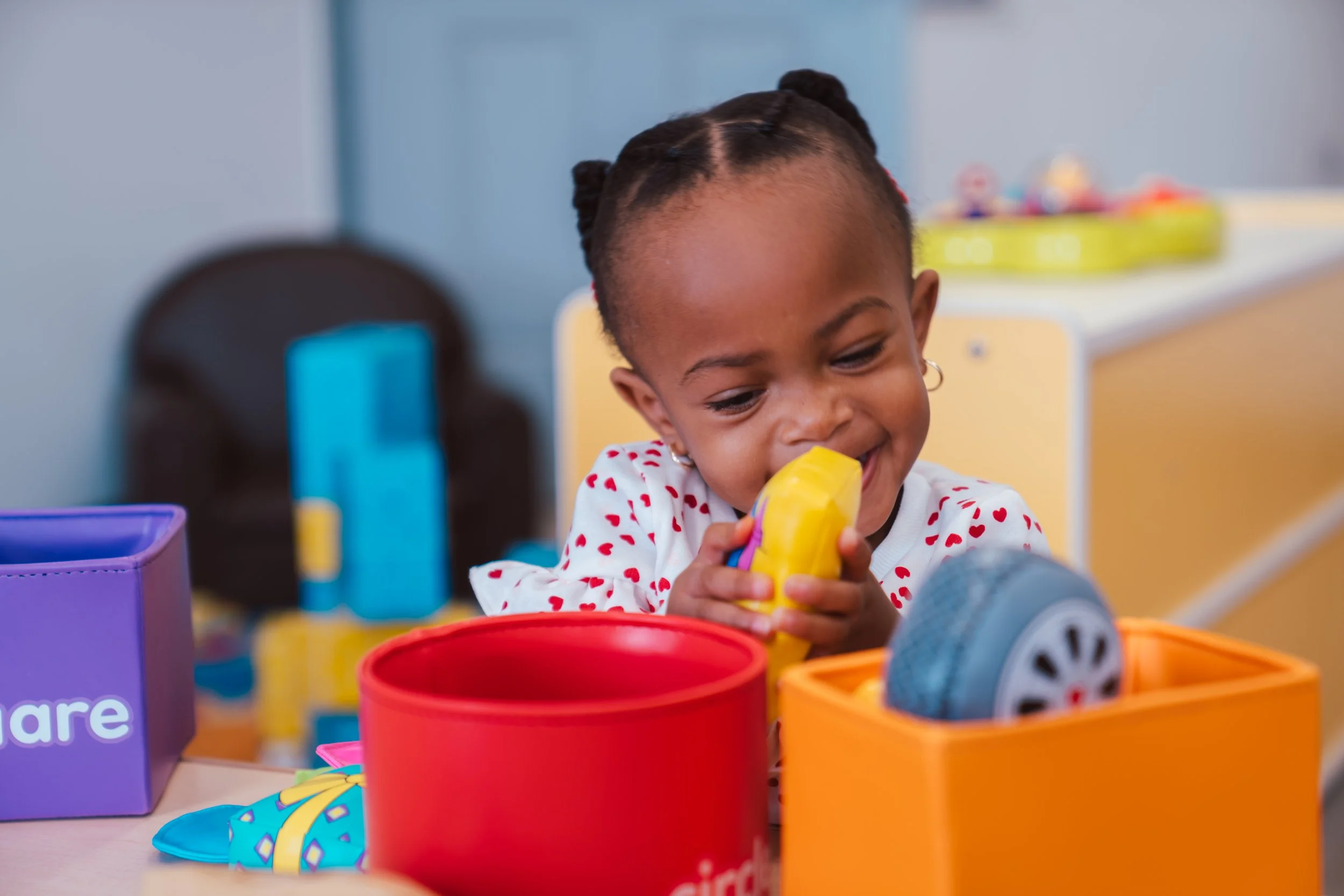 A young girl with braided hair and hoop earrings is playing with colorful toy blocks and accessories at a table in a classroom or playroom.
