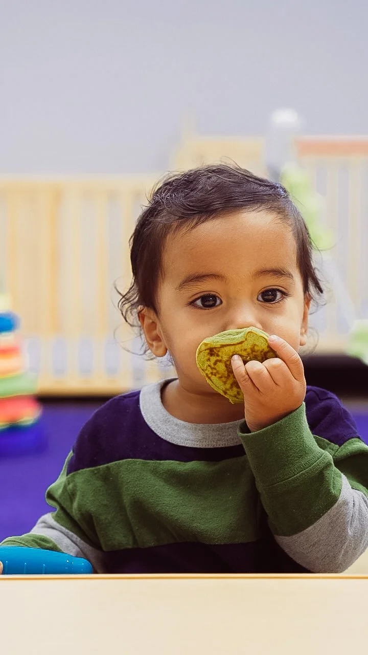 A young child with dark hair and brown eyes holding a yellow-green cookie or biscuit near his mouth, sitting at a table with a blurred colorful background.