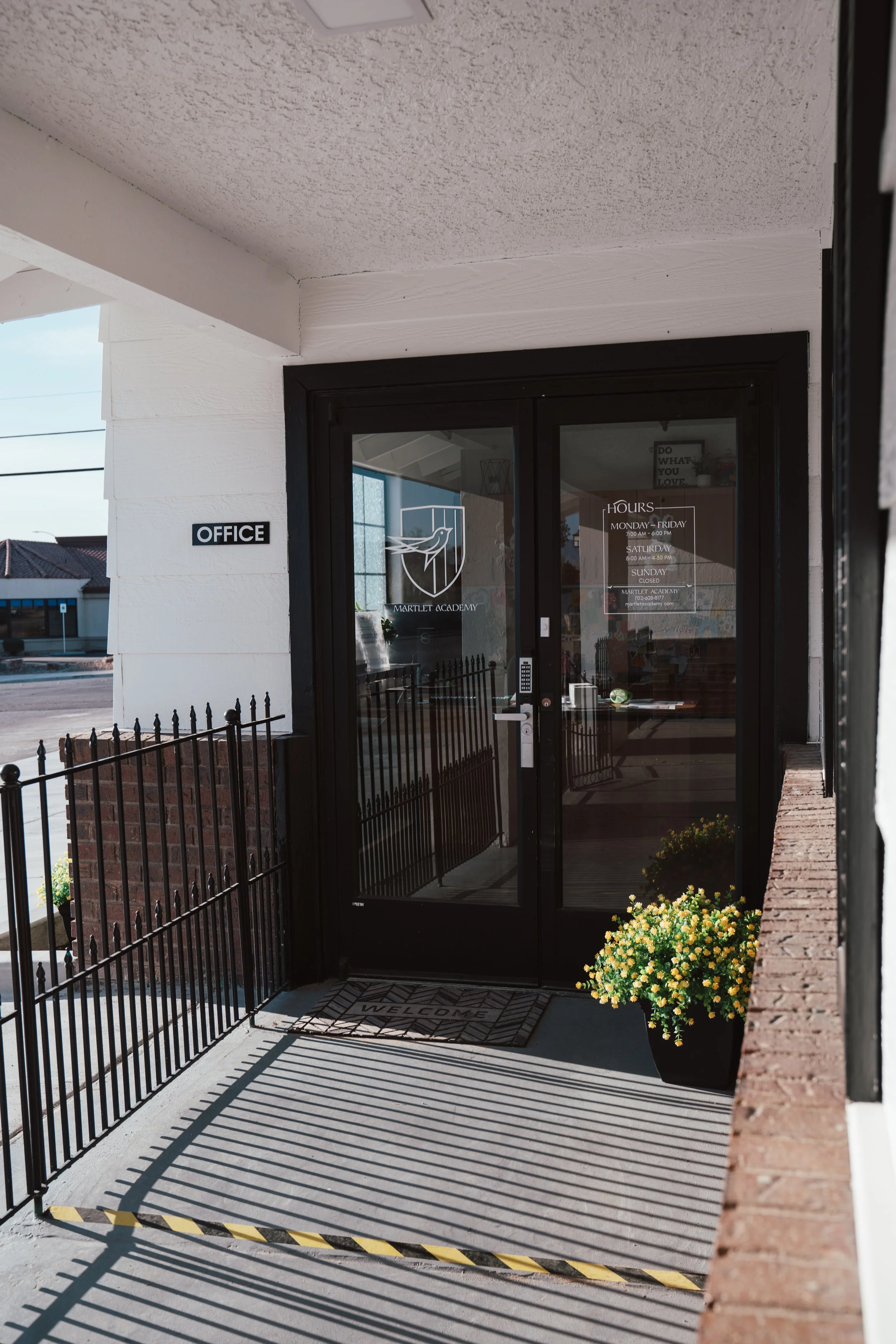 The entrance to Martlet Academy with a glass door and a black metal gate, a yellow flowering plant to the right, a welcome mat, and a sign on the door displaying hours of operation.