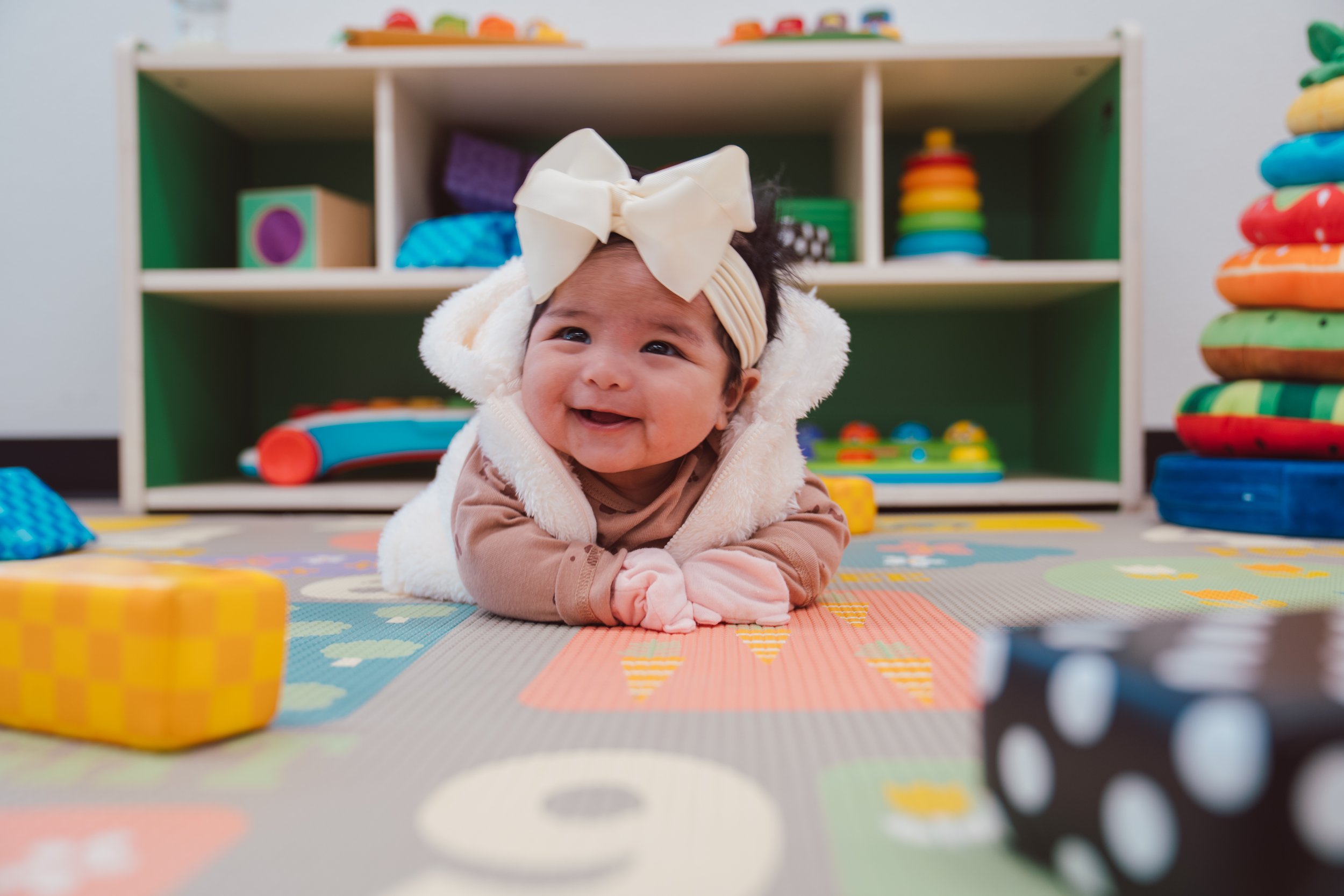 A smiling baby lying on their stomach on a colorful play mat with toys in the background.