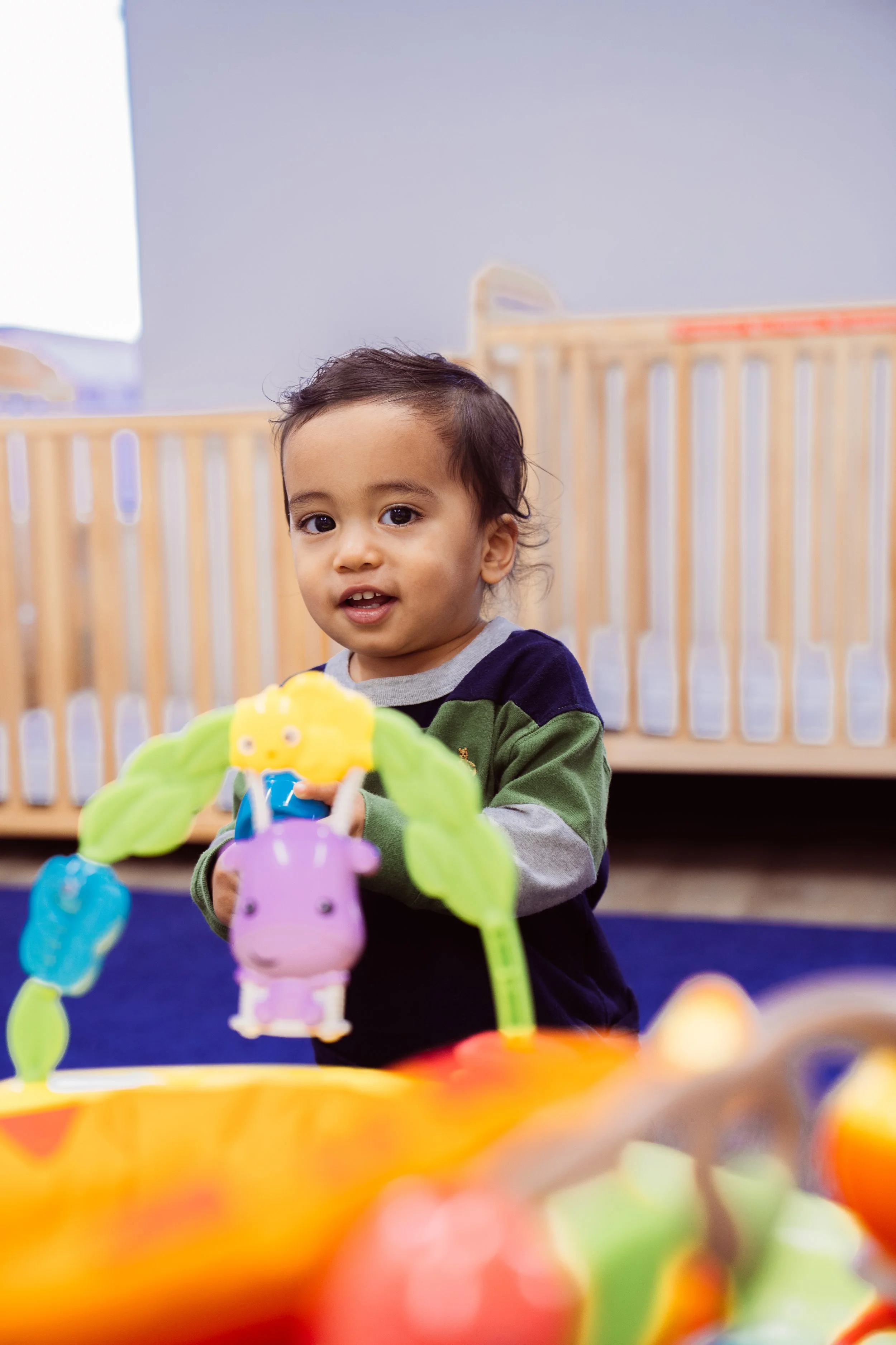A young boy with dark hair and brown eyes playing with colorful toys in a room with wooden cribs or play pens in the background.
