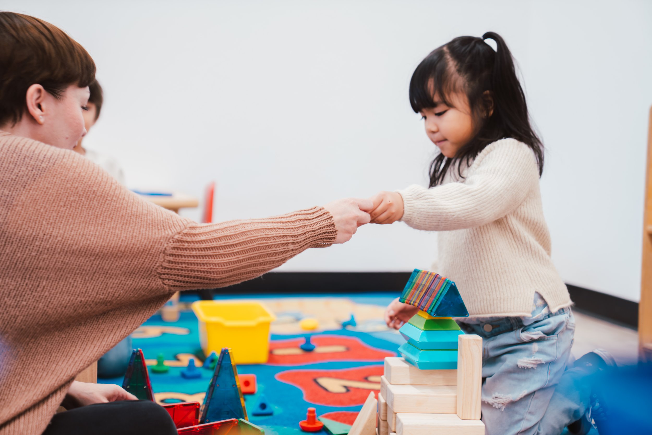 A young girl in a cream sweater and ripped jeans is playing with a woman, possibly her teacher or caregiver, at a play area with colorful blocks and toys. The woman, wearing a brownish sweater, is holding the girl's hand as they engage in an activity together.
