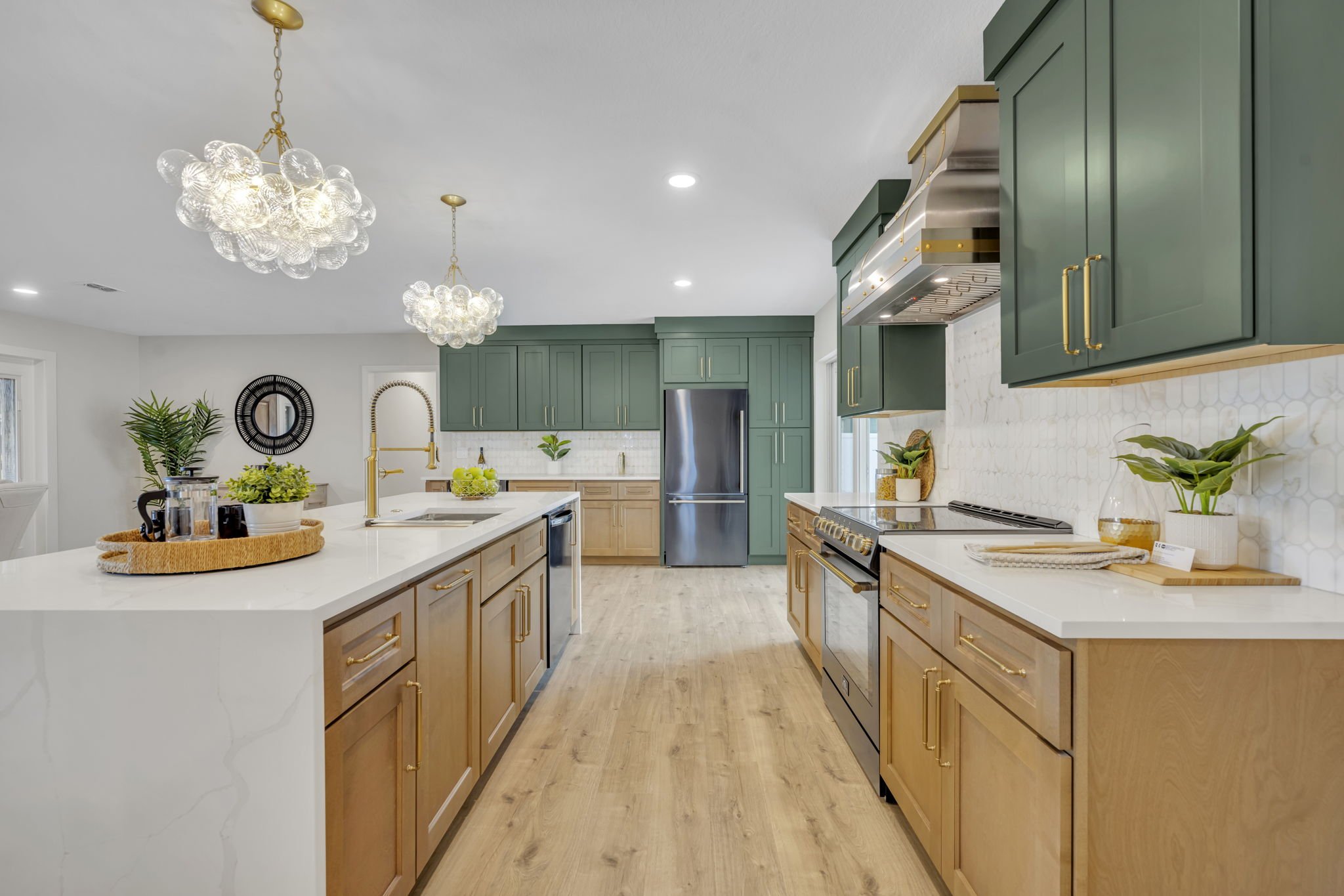 Modern kitchen with wooden and green cabinetry, a large white island with a gold faucet, and decorative plants, with pendant lights and a stainless steel refrigerator.