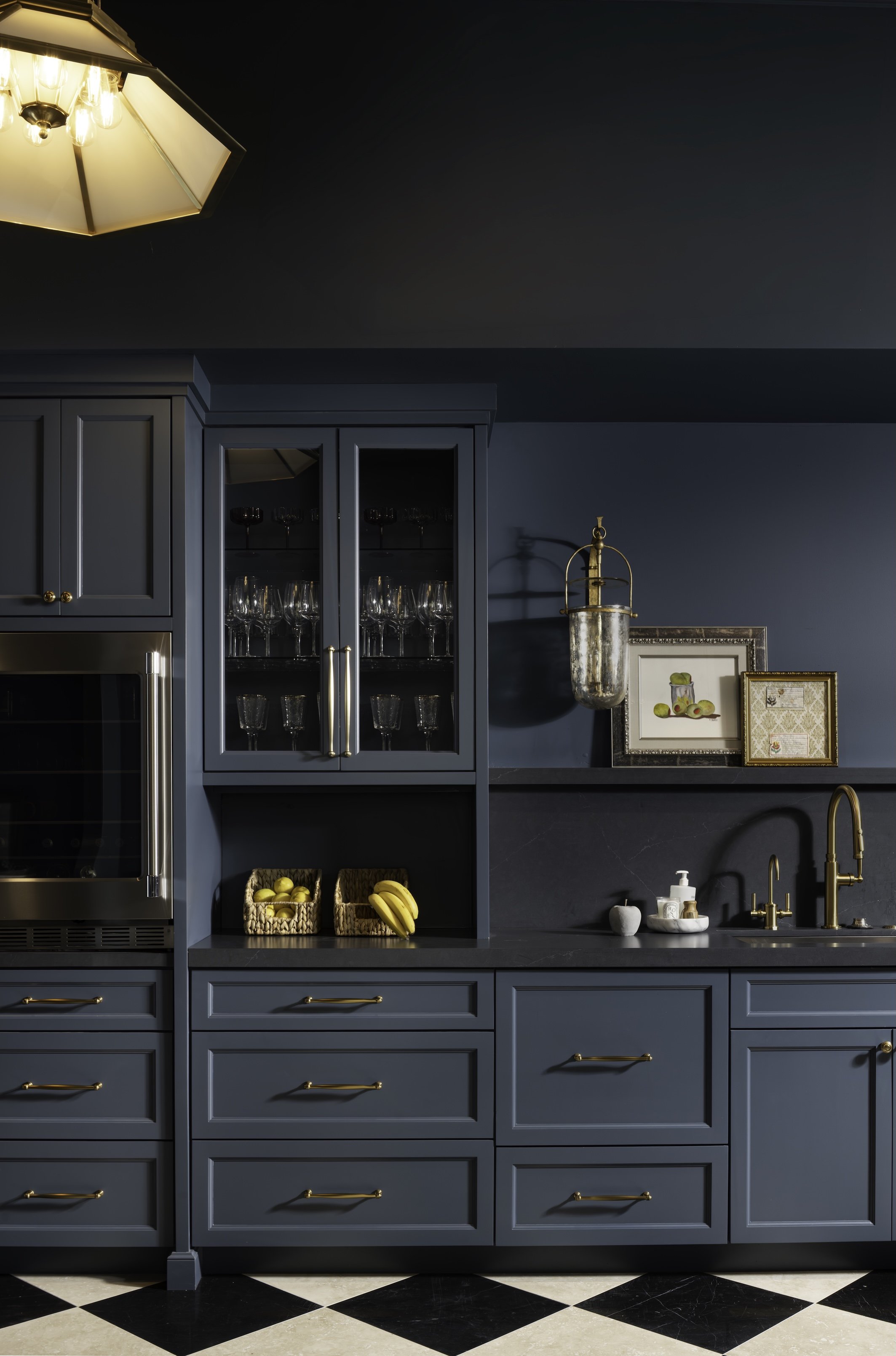 Dark blue kitchen cabinets with gold handles, glass-front cabinet with wine glasses, a basket of bananas and lemons, framed pictures, gold sink and faucet, black and white checkered floor.