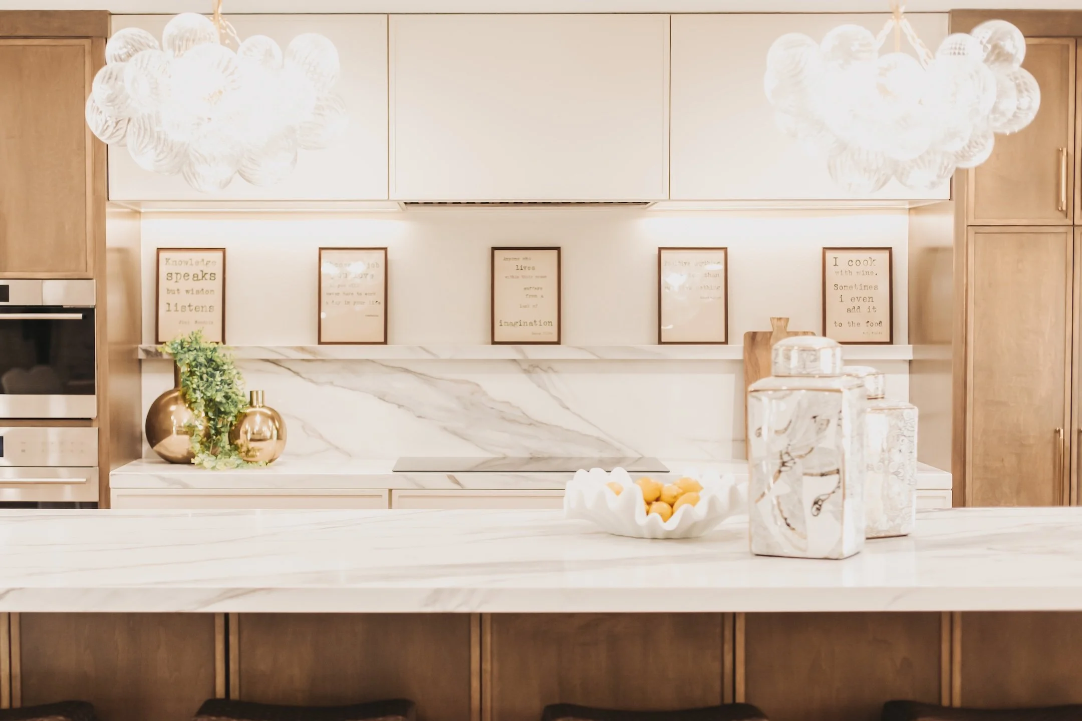 Modern kitchen with wooden cabinets, marble backsplash, and white marble countertop. Decorative items include gold vases with greenery, framed quotes on the wall, and jars on the counter.