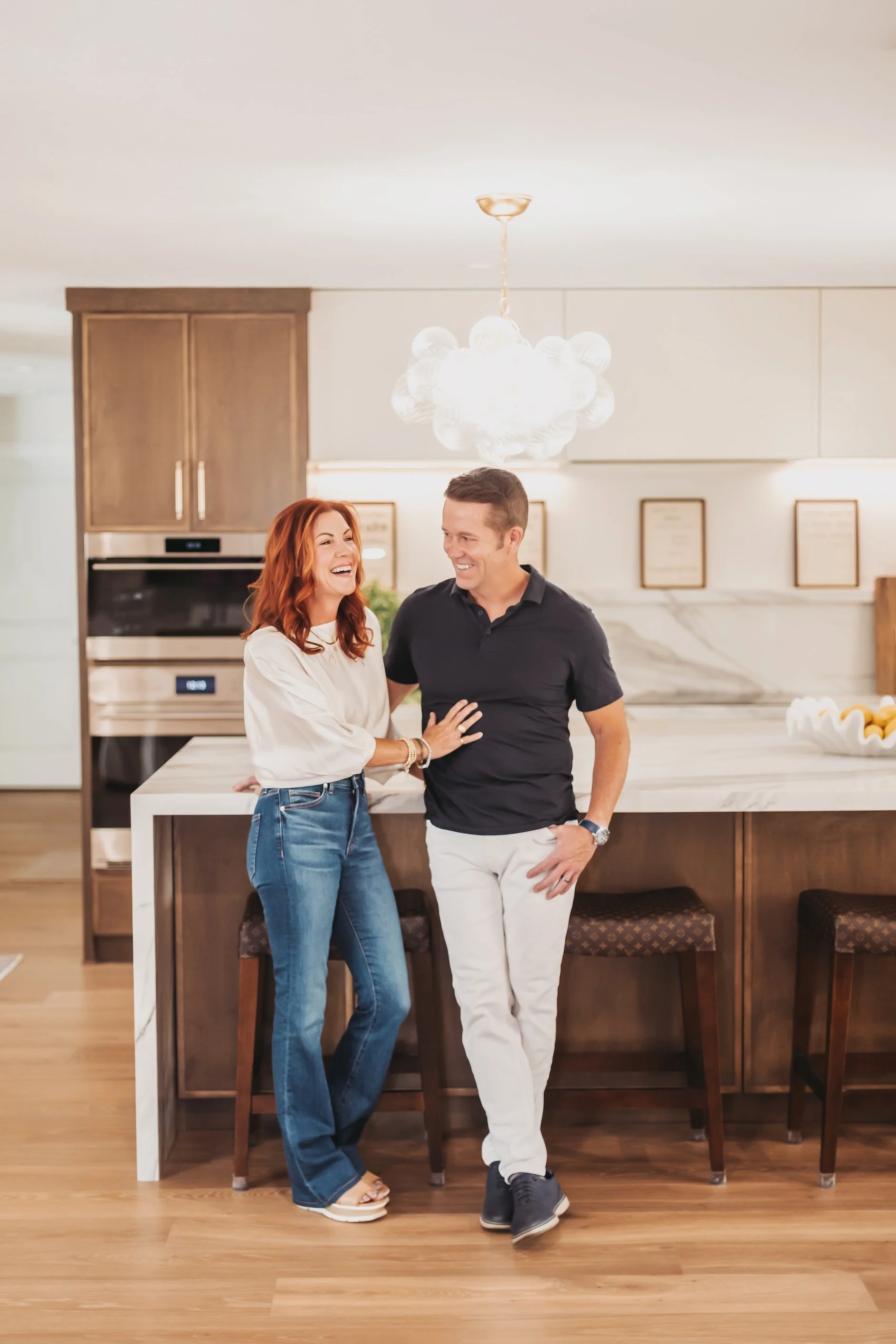 A woman with red hair and a man with short dark hair smiling and laughing together in a modern kitchen, standing near a white marble island, with wood cabinets and a hanging light fixture above.