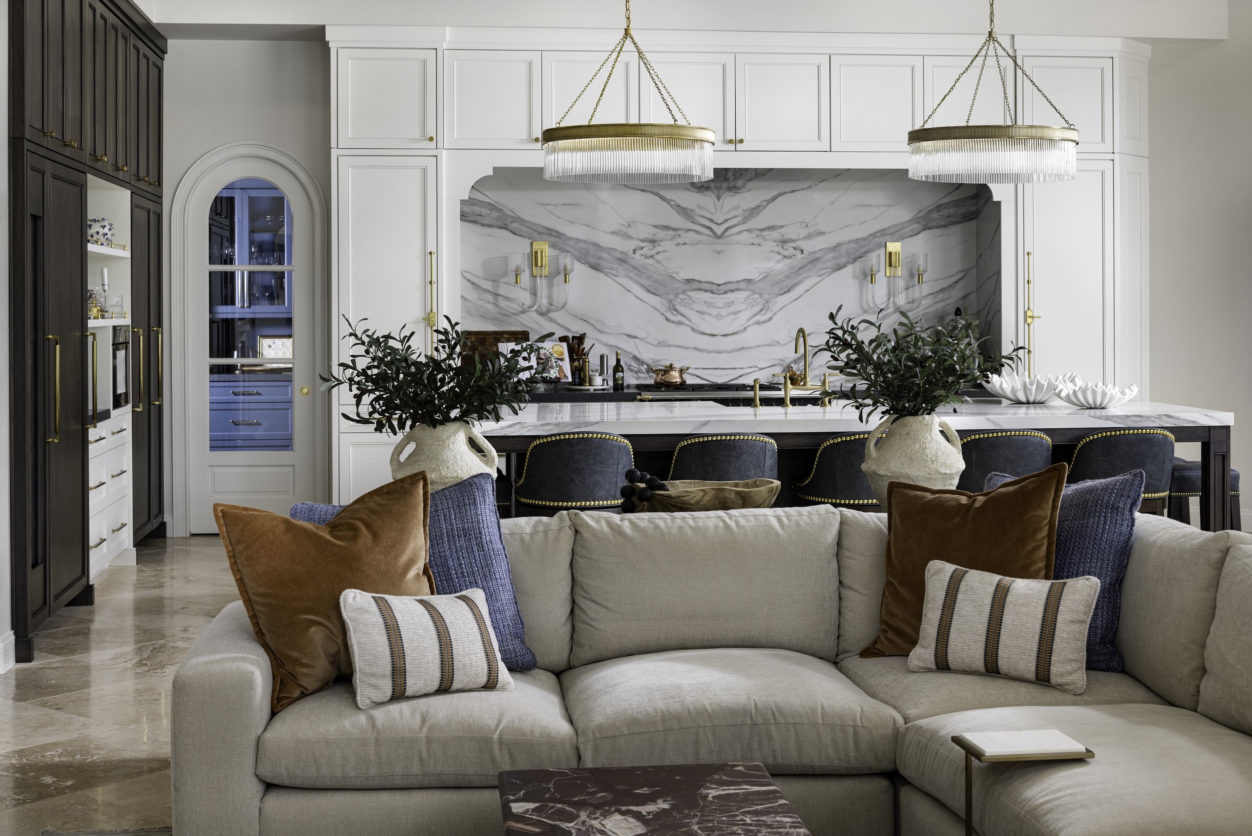 Living room with a beige sectional sofa adorned with brown, navy, and striped pillows, facing a kitchen with white and dark cabinetry, marble backsplash, and gold accents, including two chandeliers.