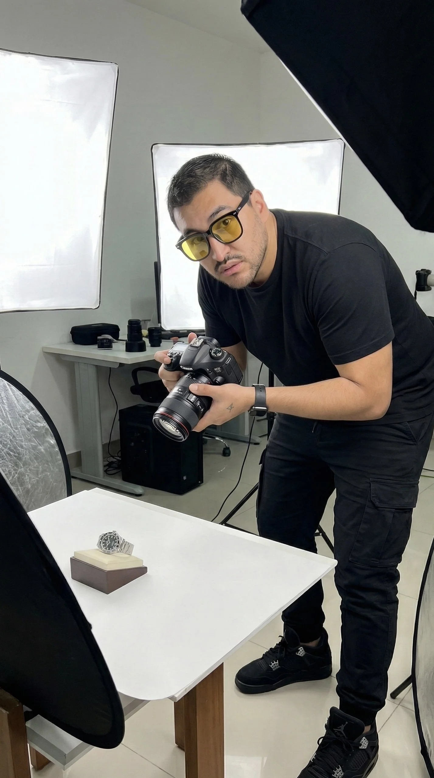 Hombre con gafas de sol sosteniendo una cámara en un estudio de fotografía, en primer plano, con una caja de reloj en una mesa y equipos de iluminación en el fondo.