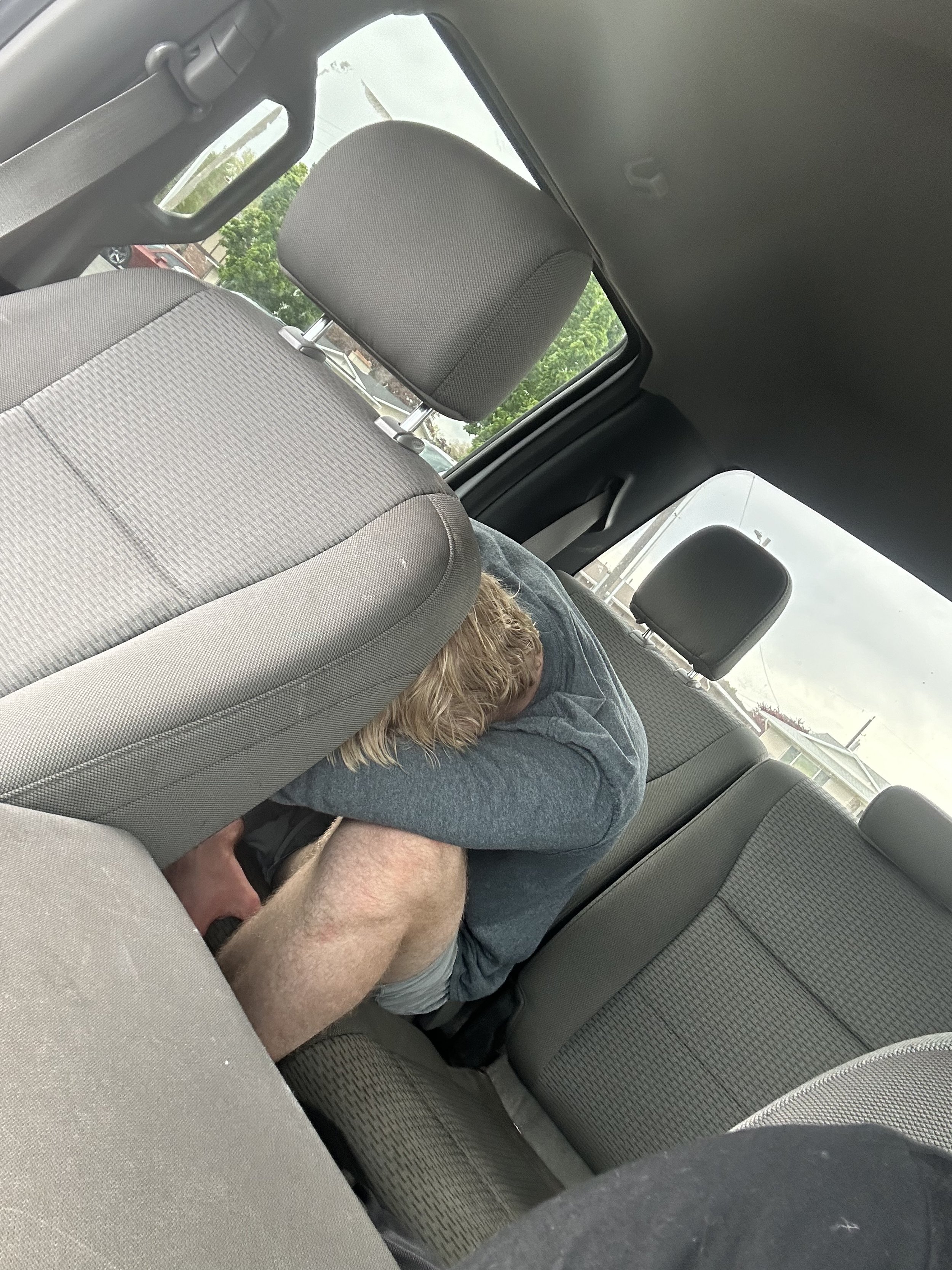 A person with blond hair, wearing a gray hoodie and shorts, is sitting in the back seat of a car, leaning forward with their head resting on the front passenger seat.