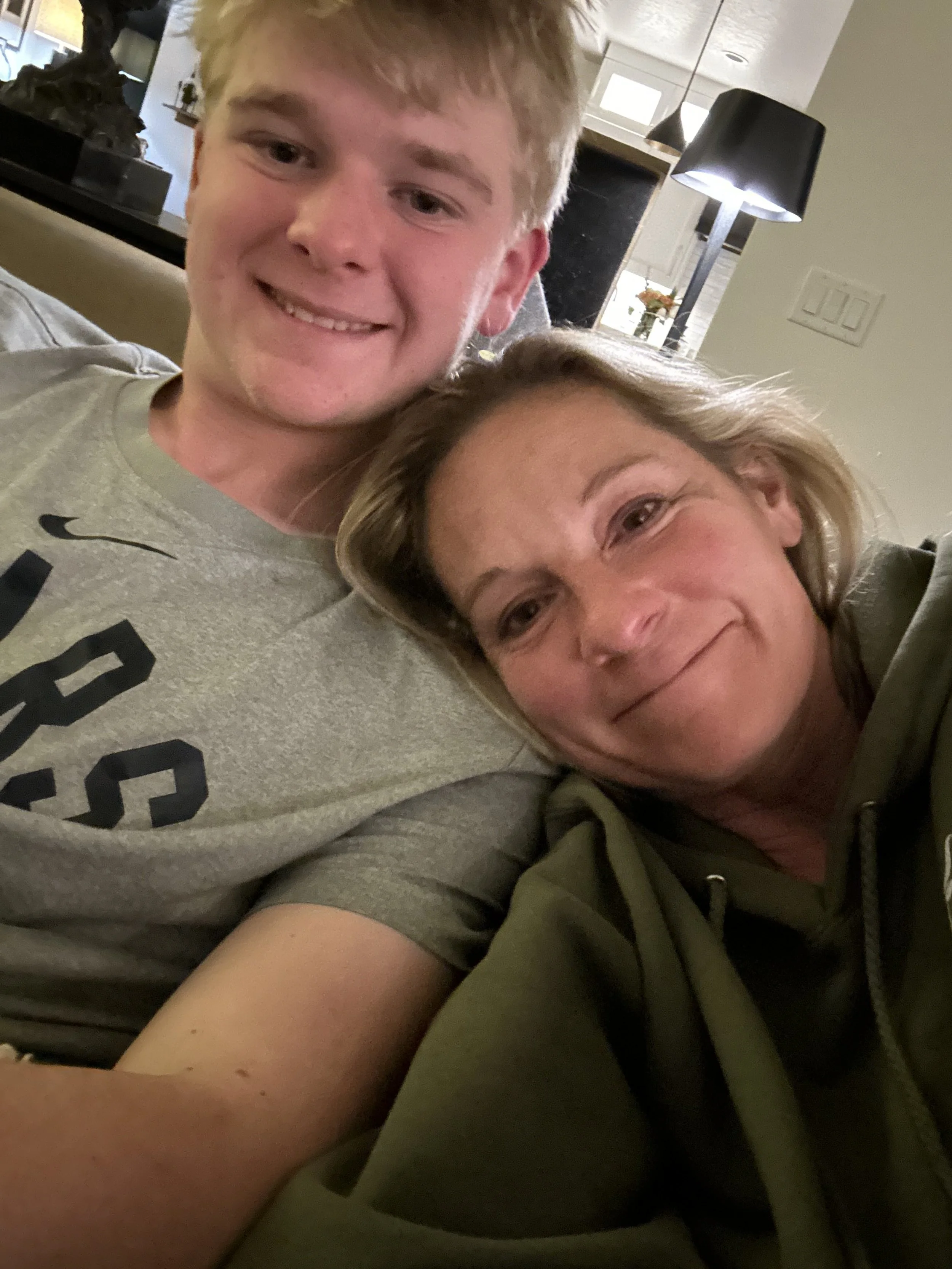 A smiling young man and an older woman sitting closely and taking a selfie together inside a house.