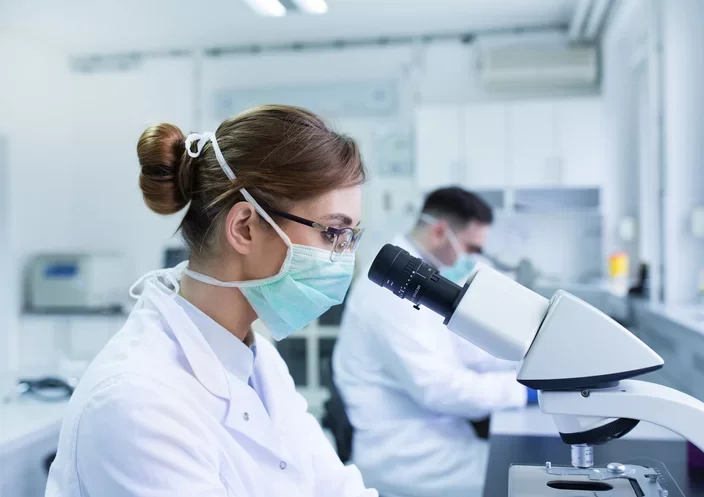 A scientist in a laboratory wearing a mask and glasses using a microscope.