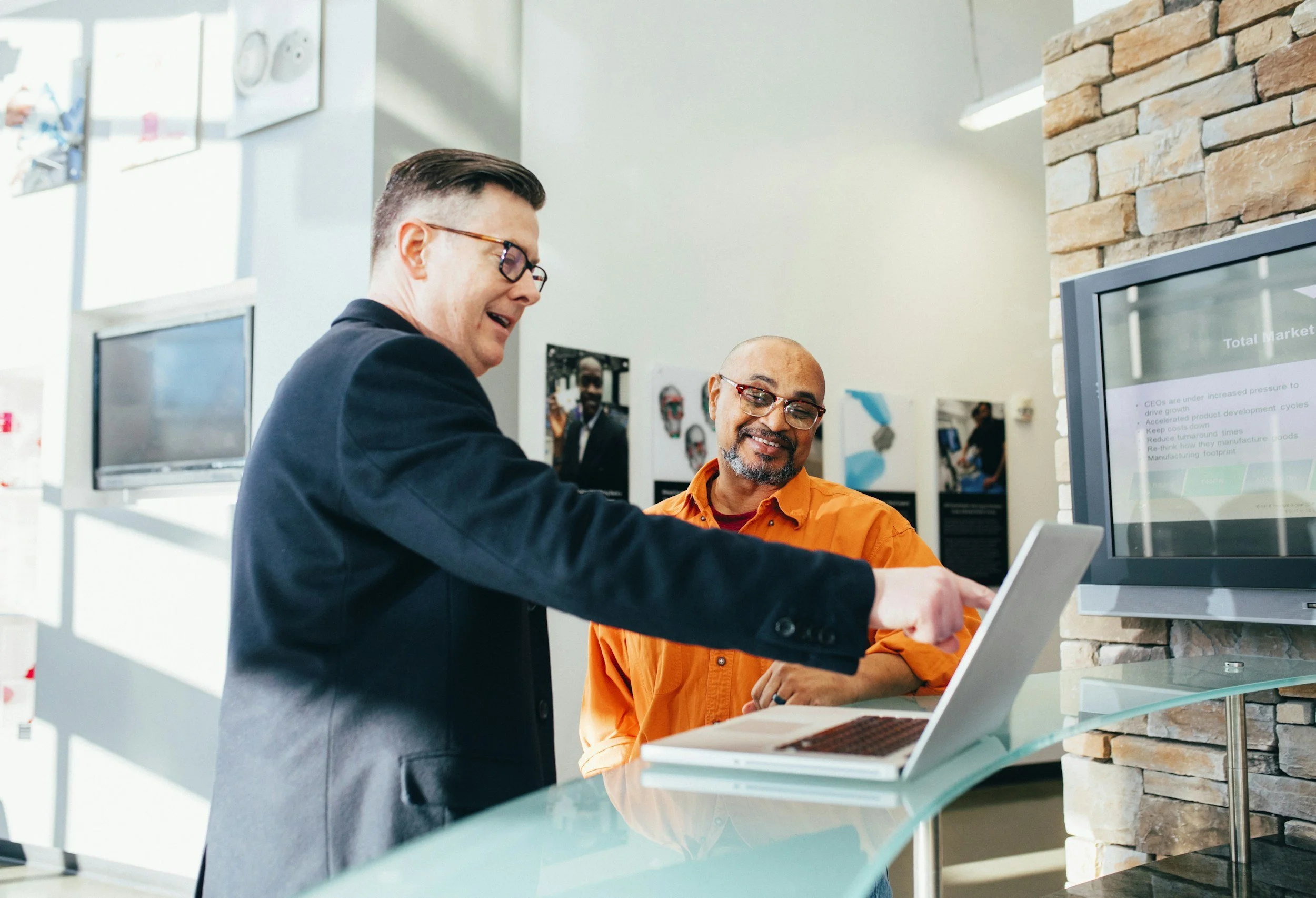 Two men are looking at a laptop computer on a glass counter. One is pointing at the screen and the other is smiling. The setting appears to be a modern office or showroom with a brick wall and multiple monitors in the background.