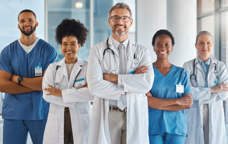 Group of five healthcare professionals including doctors and nurses standing together in a hospital hallway, smiling.