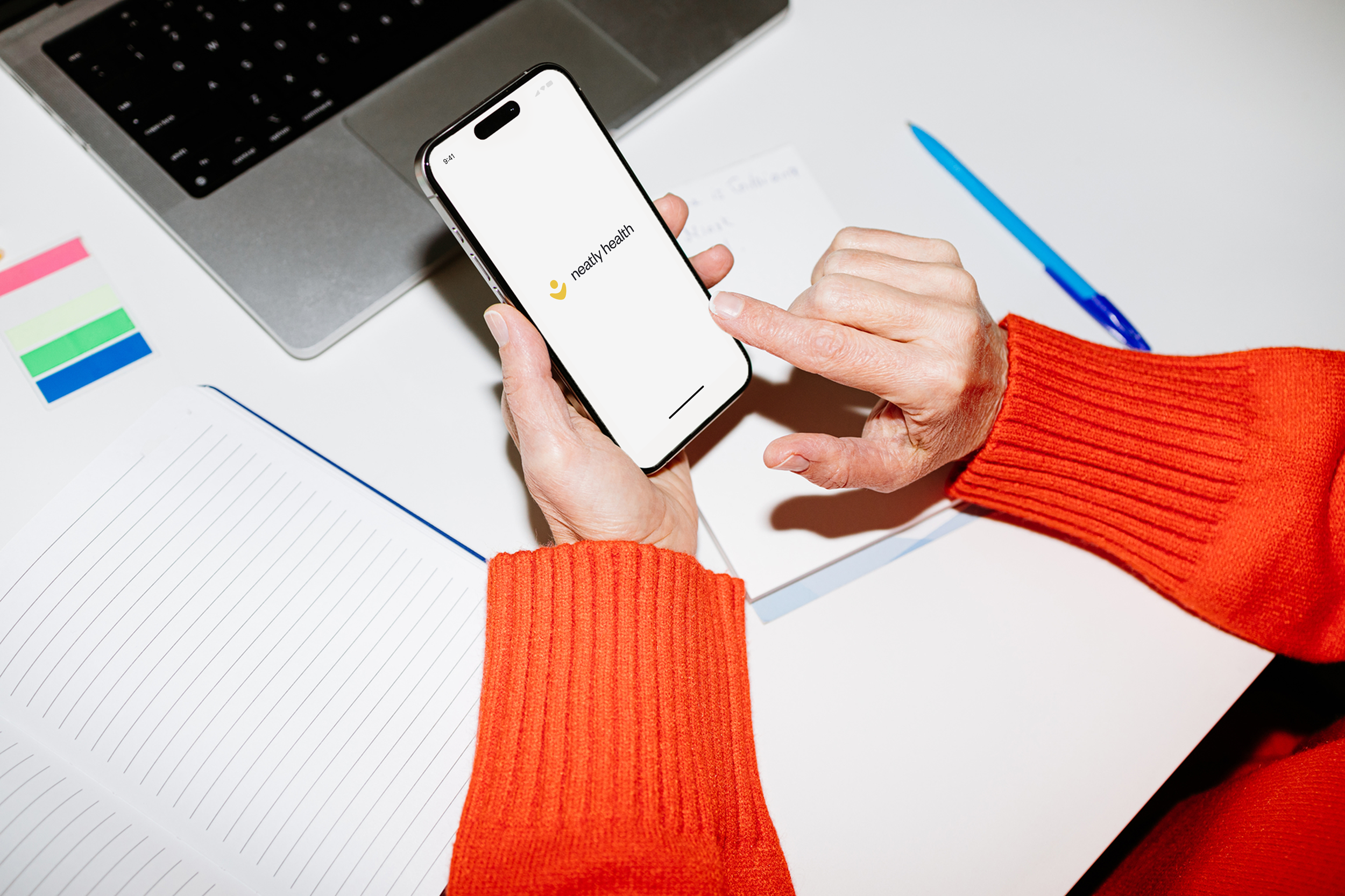 A person in a red sweater using a smartphone with the Neatly Health app open, placed on a white desk with a notebook, a blue pen, color swatches, and a laptop.