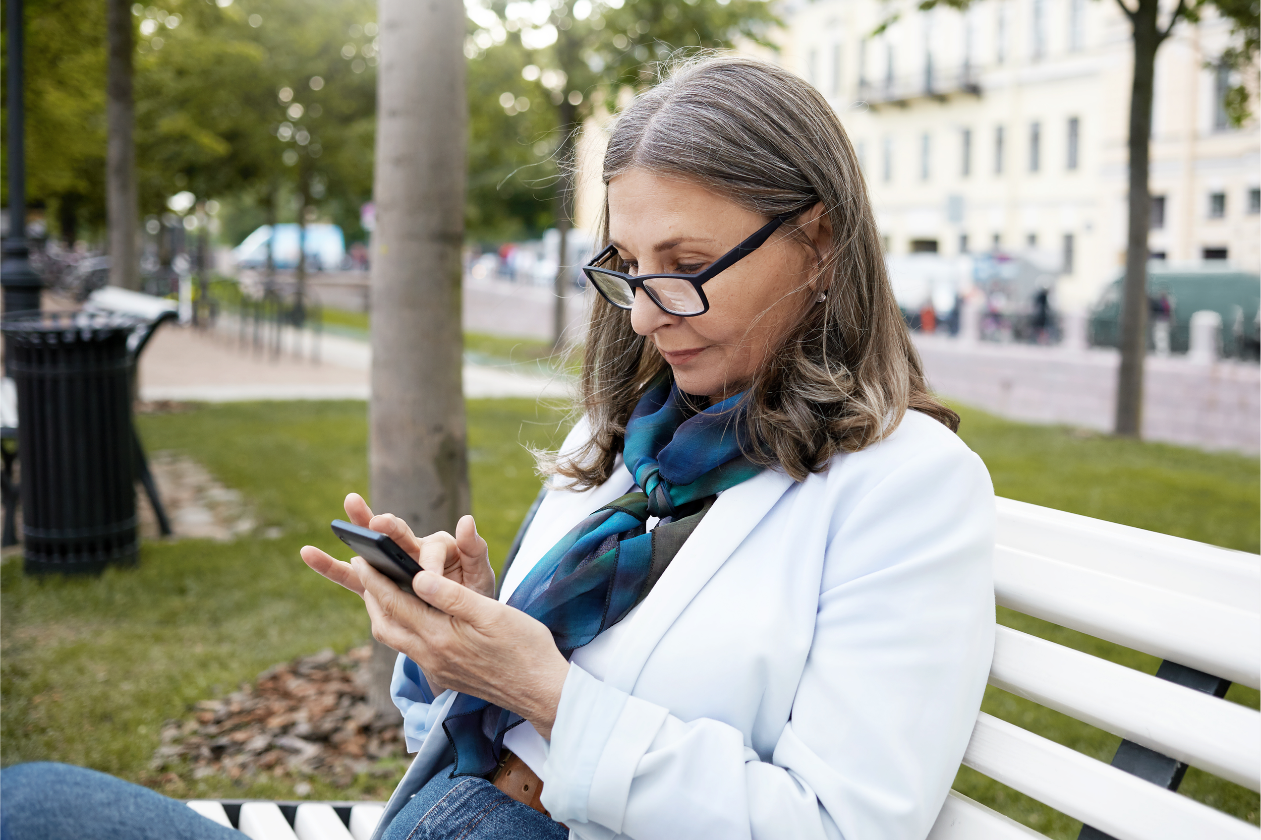 Middle-aged woman sitting on a park bench, looking at her smartphone, outdoors with trees and city buildings in the background.