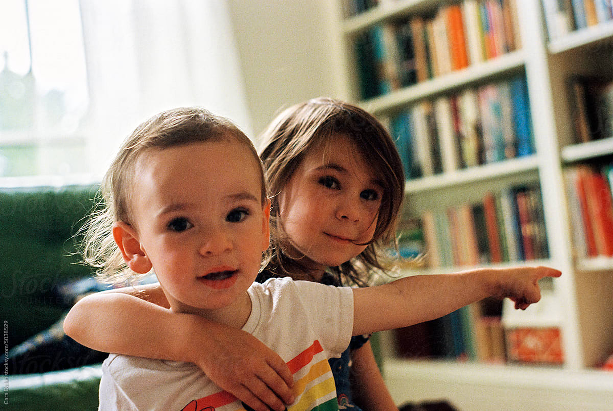 Two children, a boy with short hair and a girl with longer hair, sitting together in a room with a bookshelf filled with colorful books in the background. The girl points to the right while the boy looks toward the camera.