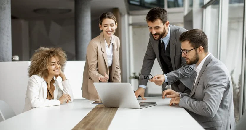 Four businesspeople in a meeting, two men and two women, are gathered around a table with a laptop, engaging in a discussion and smiling.