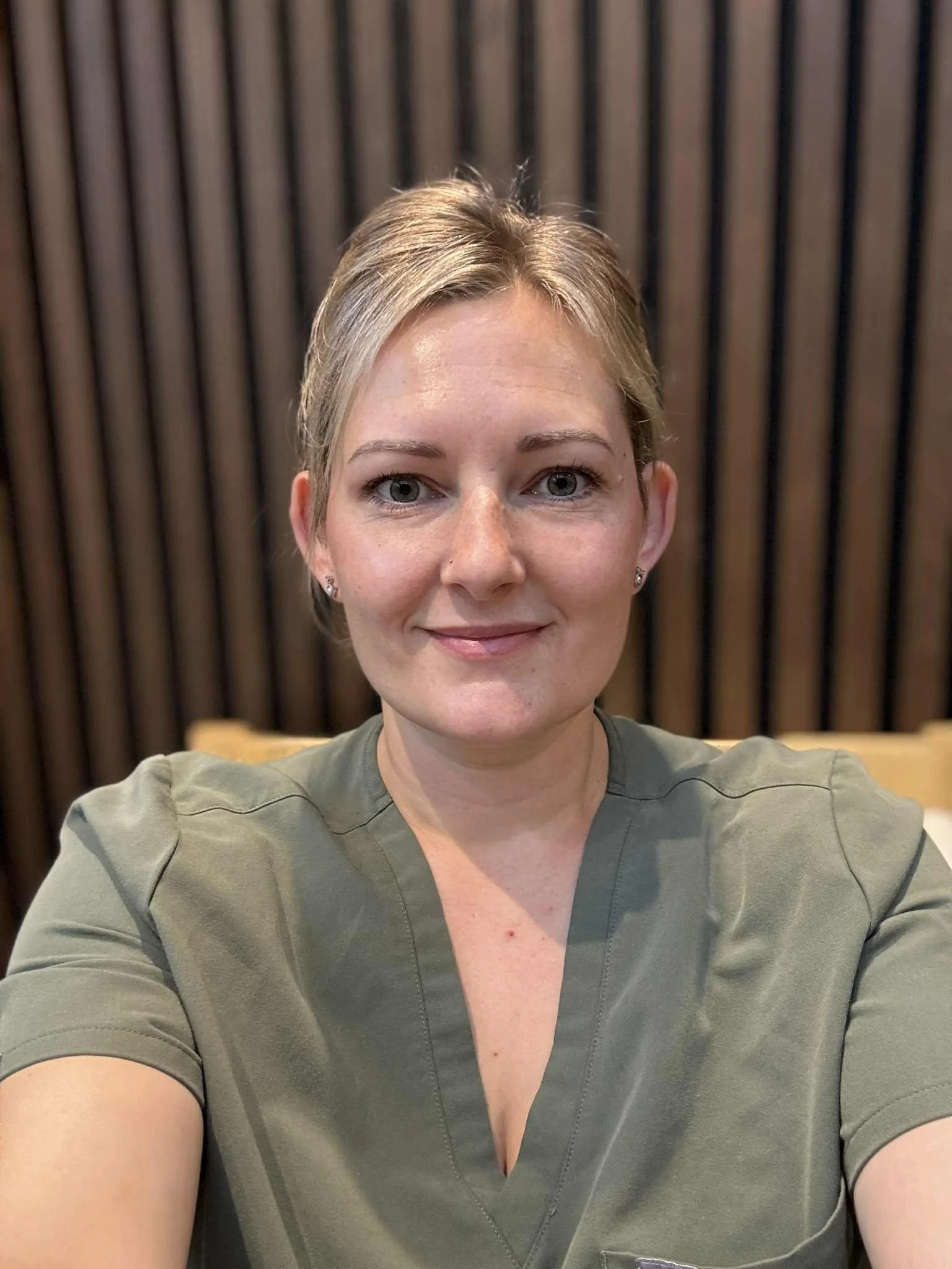 A woman taking a selfie in front of a dark wooden paneled wall, wearing a green short-sleeved top