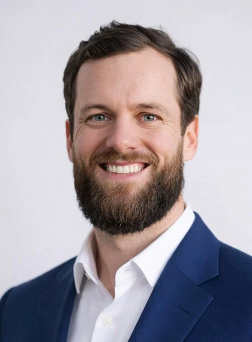A man with a beard and short brown hair, wearing a dark blue suit jacket and white shirt, smiling against a plain white background.